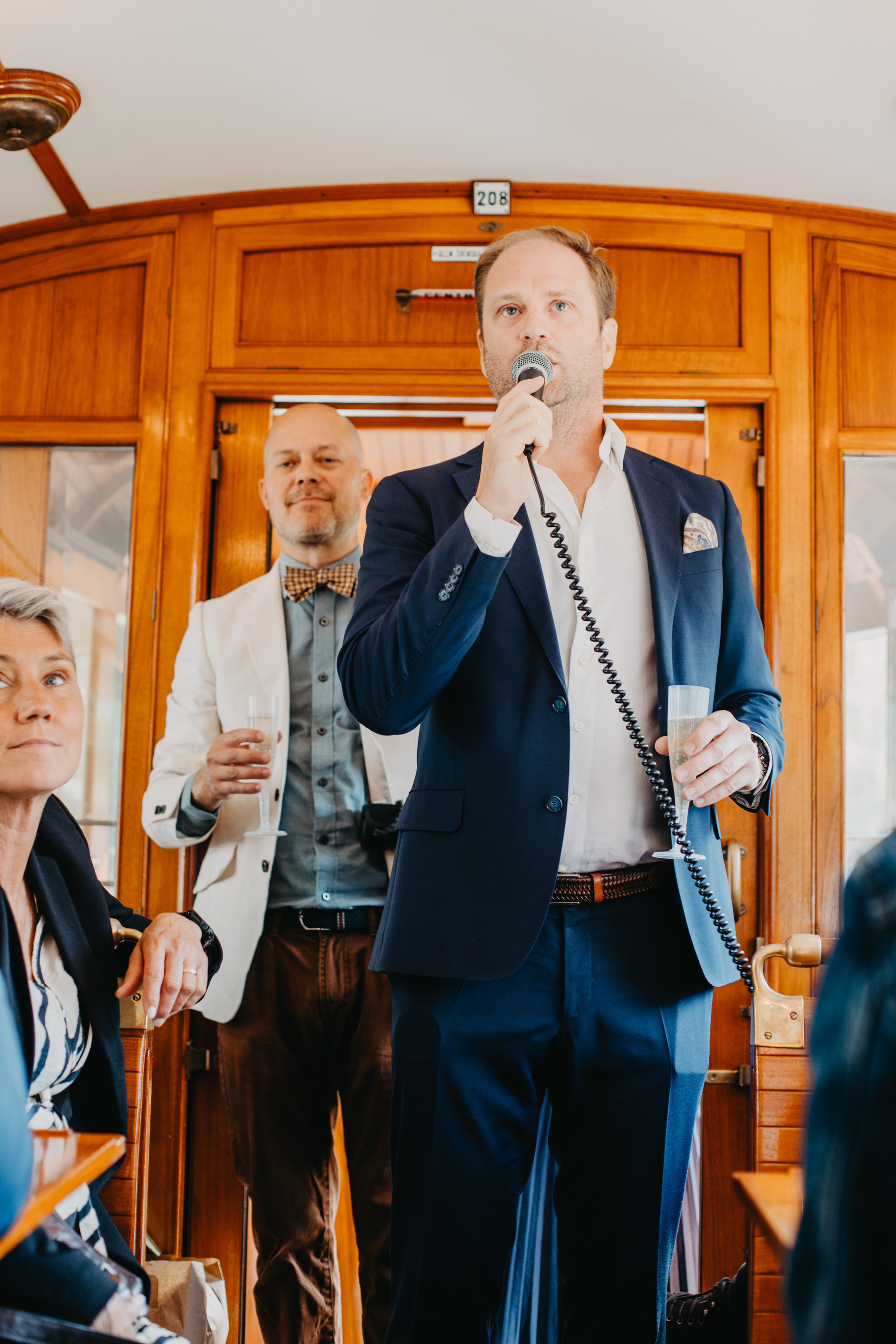 Auto-generated description: A man in a dark suit is speaking into a microphone inside a wooden-paneled room, while another man in a white jacket stands behind him holding a drink.