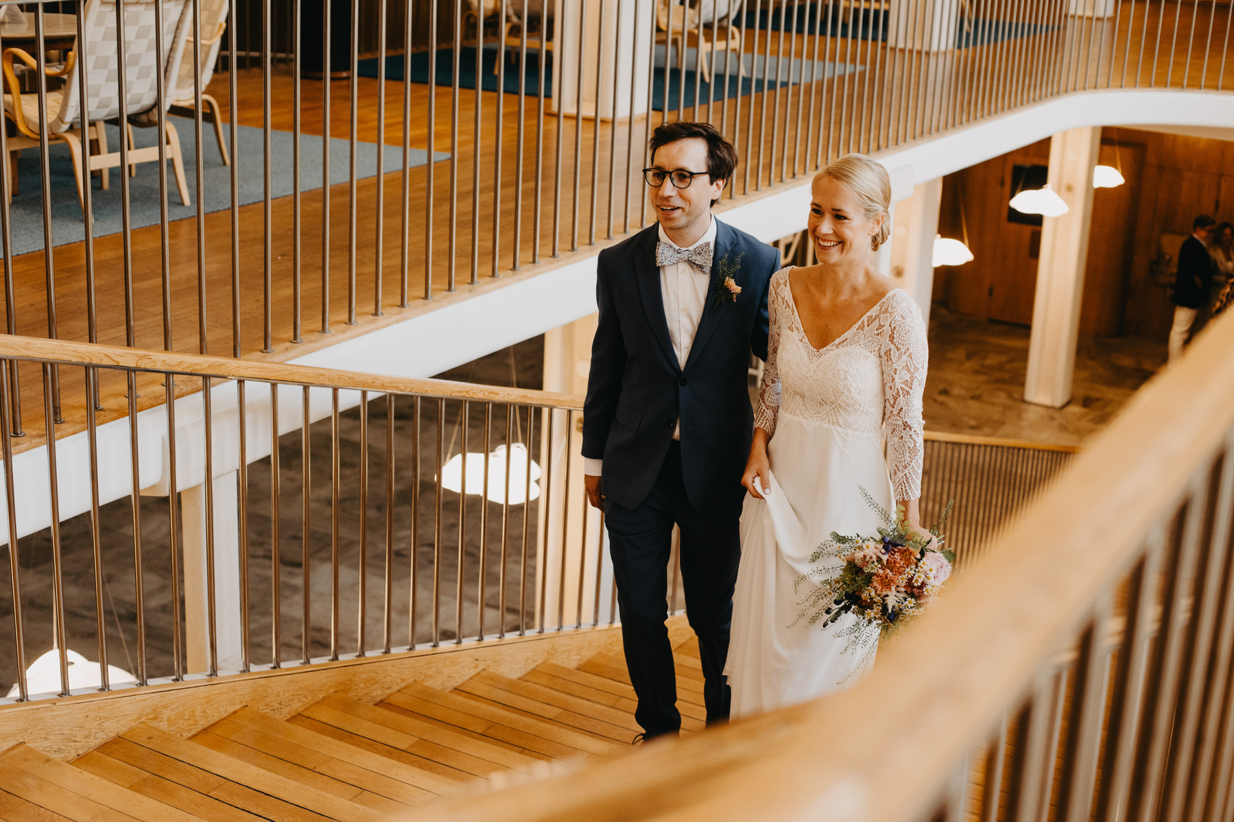 Auto-generated description: A couple dressed in wedding attire is walking up a wooden staircase, with the bride holding a bouquet of flowers.