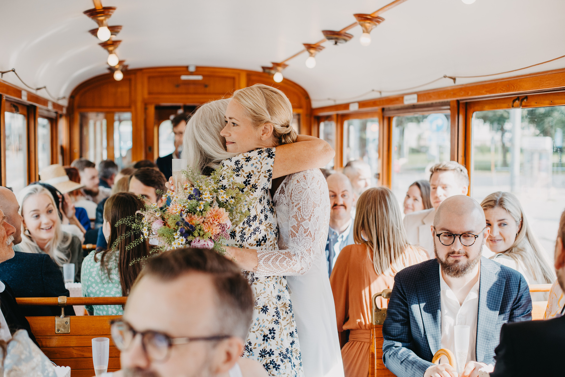 Auto-generated description: Two women embrace affectionately in a warmly lit train carriage surrounded by smiling people.