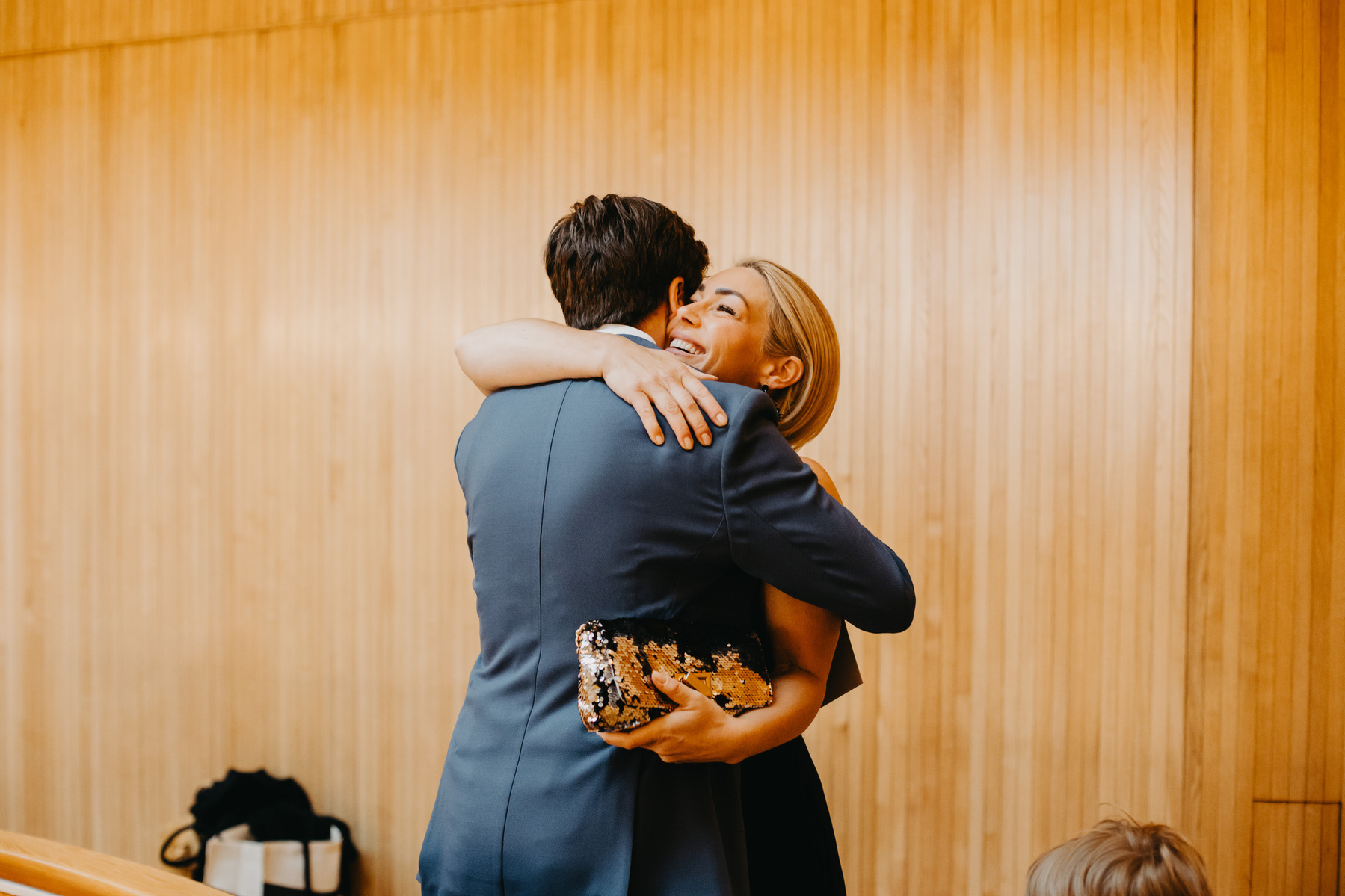 Auto-generated description: Two people are hugging warmly in front of a wooden wall, with one holding a clutch bag.