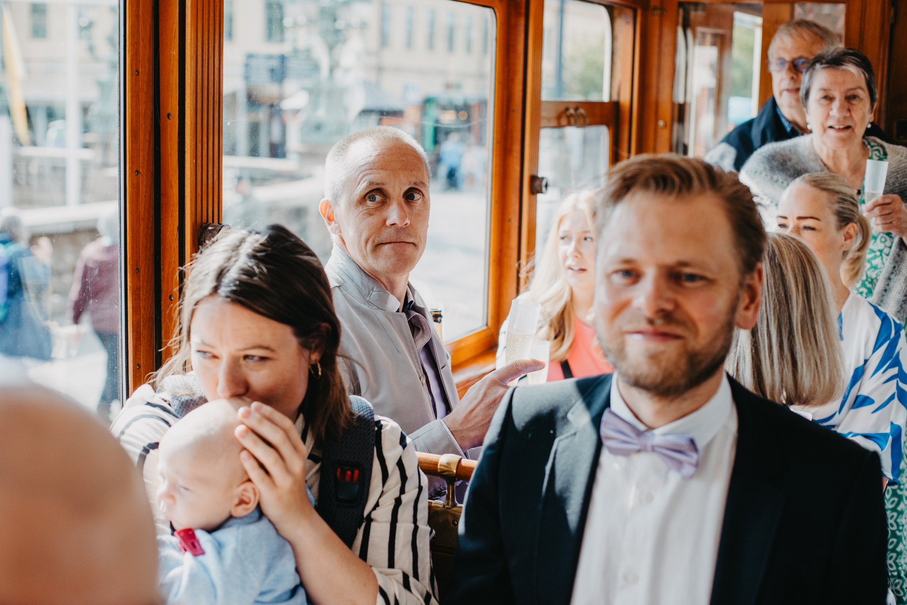 Auto-generated description: A group of people, including a woman holding a baby and a man in a bow tie, are gathered inside what appears to be a tram or bus.