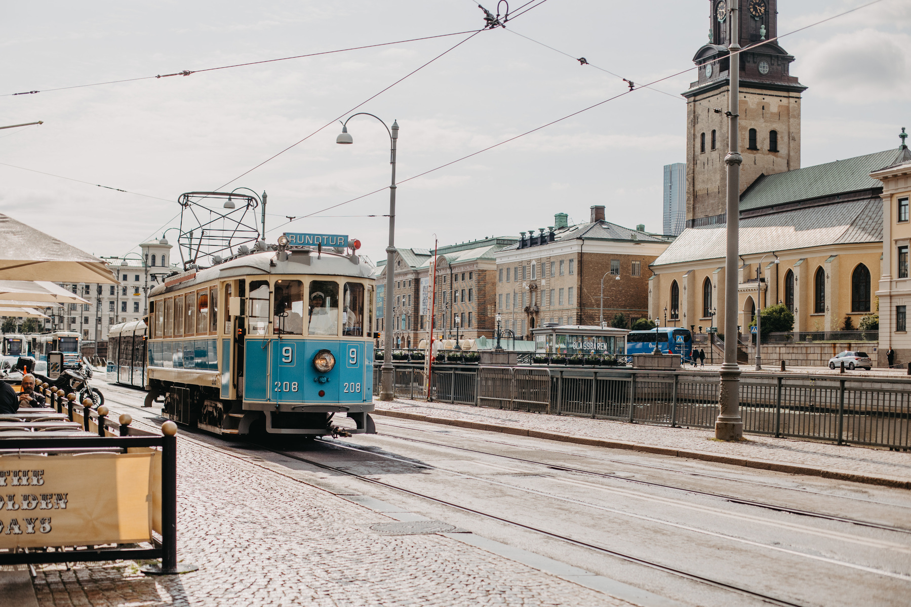 Auto-generated description: A blue and white tram is traveling through a historic European city street with a clock tower in the background.