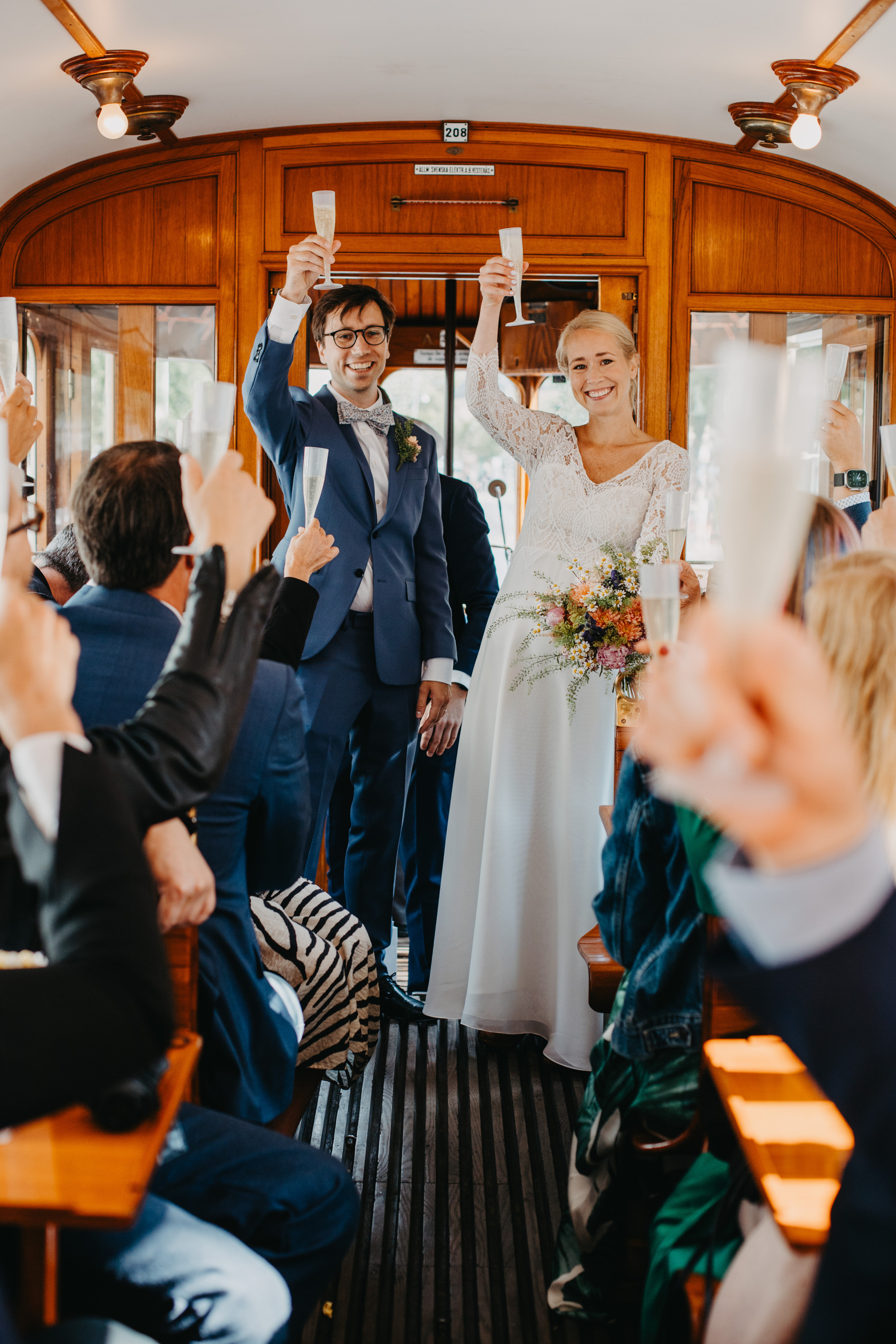 Auto-generated description: A smiling bride and groom in wedding attire raise champagne glasses, joined by cheering guests inside a wooden trolley.