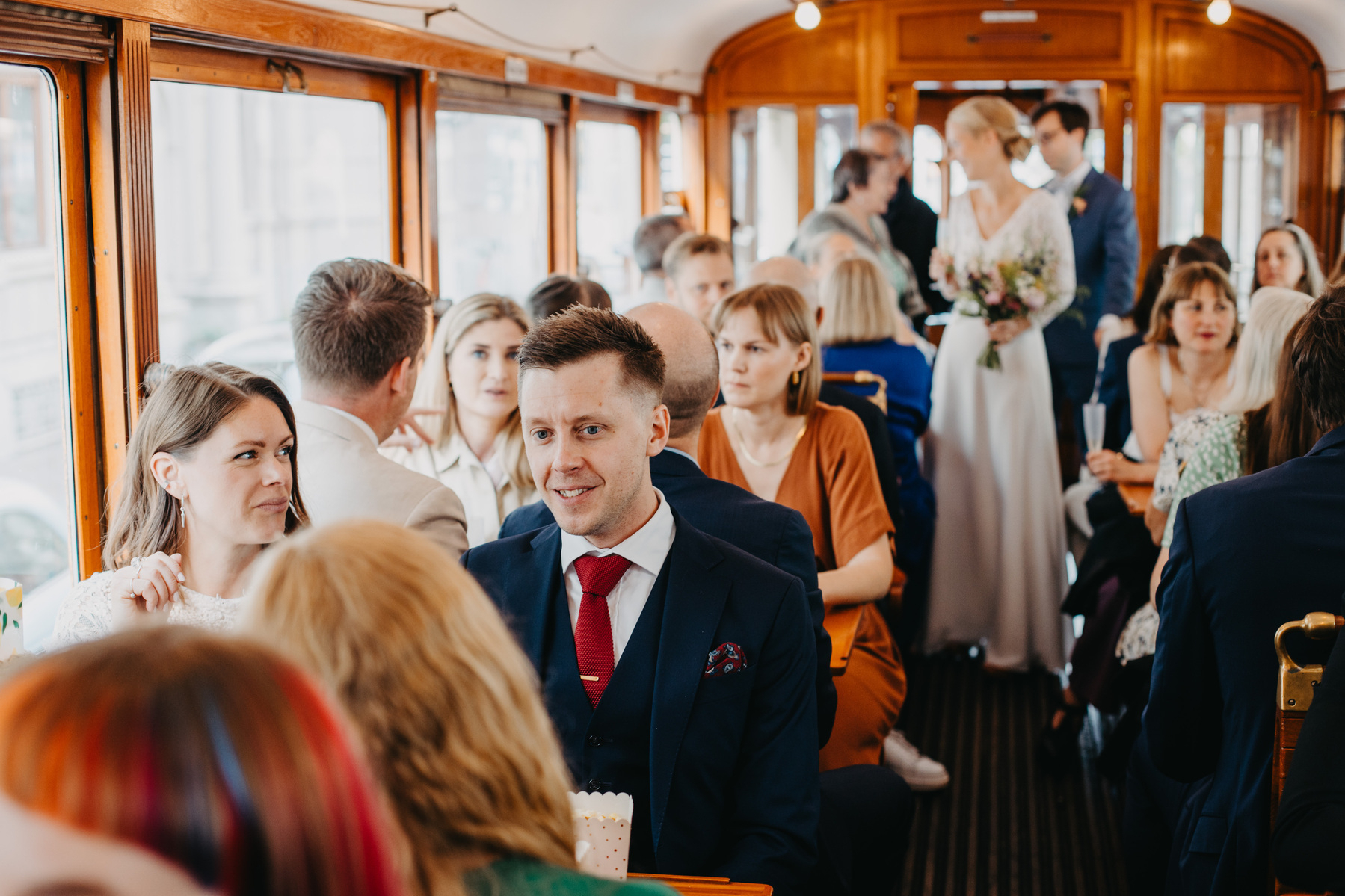 Auto-generated description: A group of people dressed in formal attire are seated and standing inside a vintage train carriage, with a couple at the far end seemingly in a wedding ceremony.