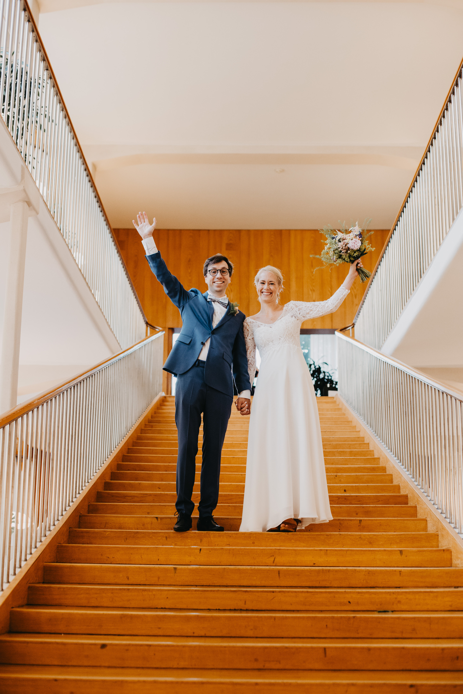 Auto-generated description: A smiling couple in wedding attire is holding hands and standing on a staircase, with both raising one arm in celebration.