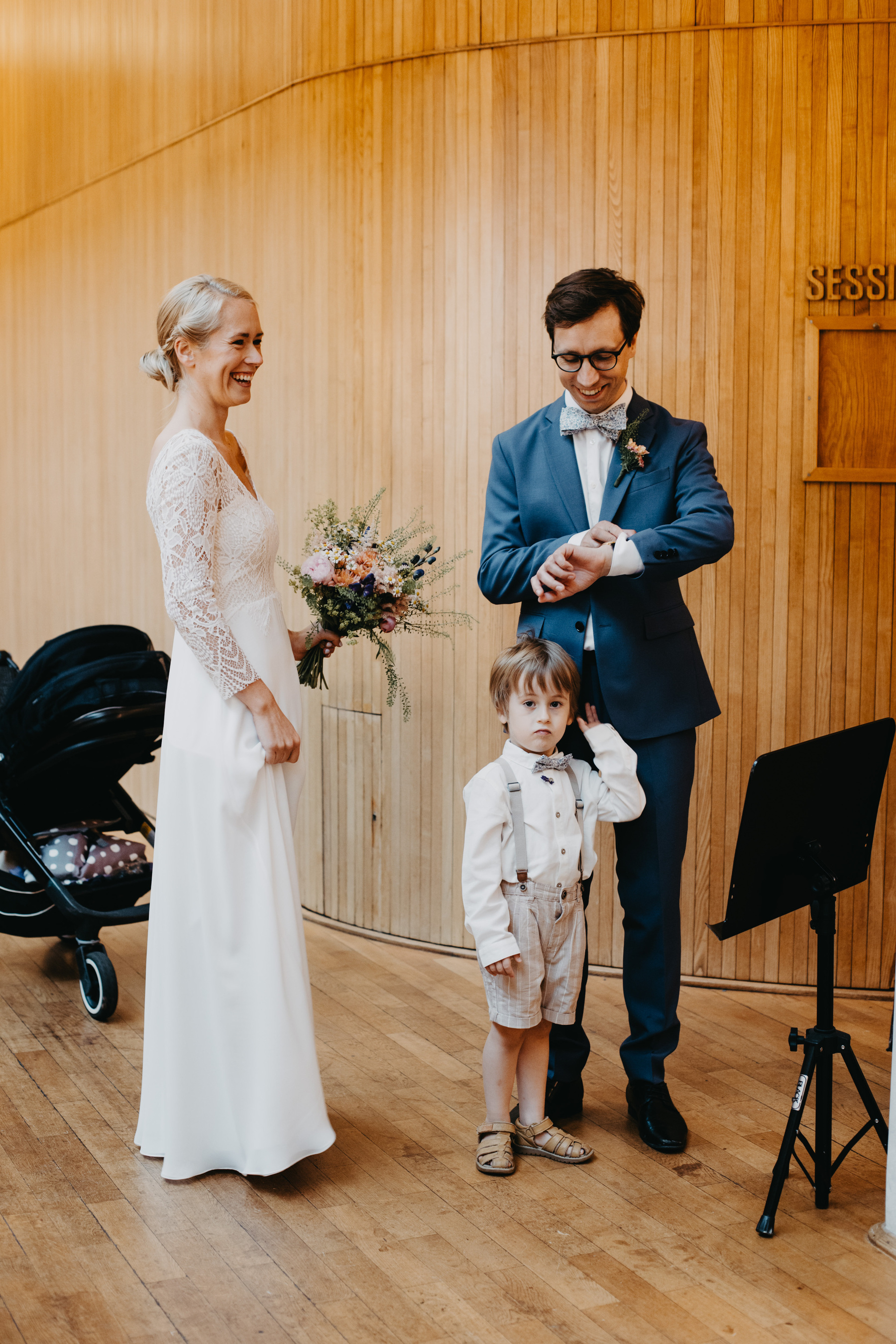 Auto-generated description: A woman in a white dress, a man in a blue suit, and a young boy stand indoors near a stroller and a music stand, with the man checking his watch while the woman smiles.