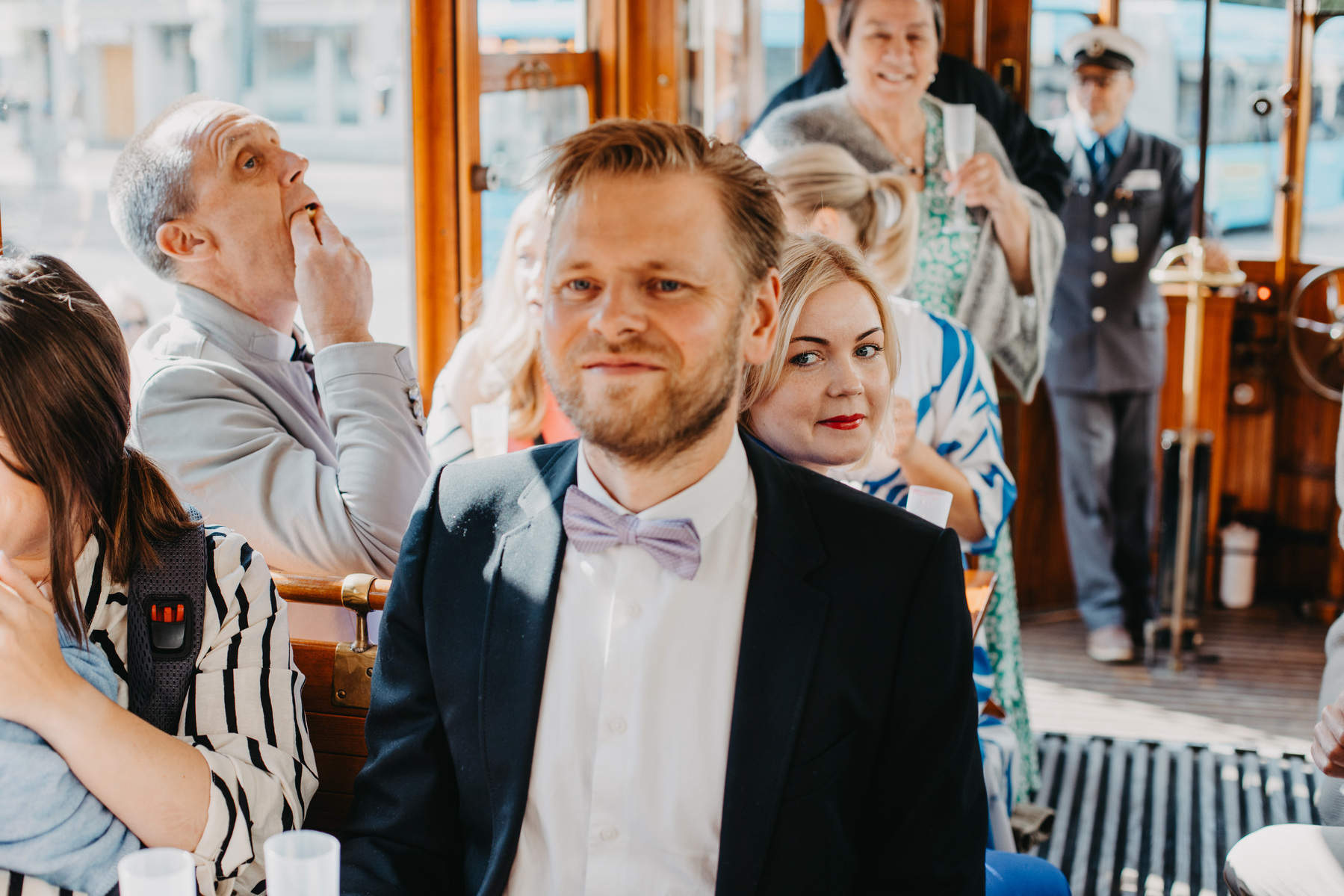 Auto-generated description: A man in a suit and bow tie sits on a bus or tram with several other passengers, some of whom are looking around or at the camera.