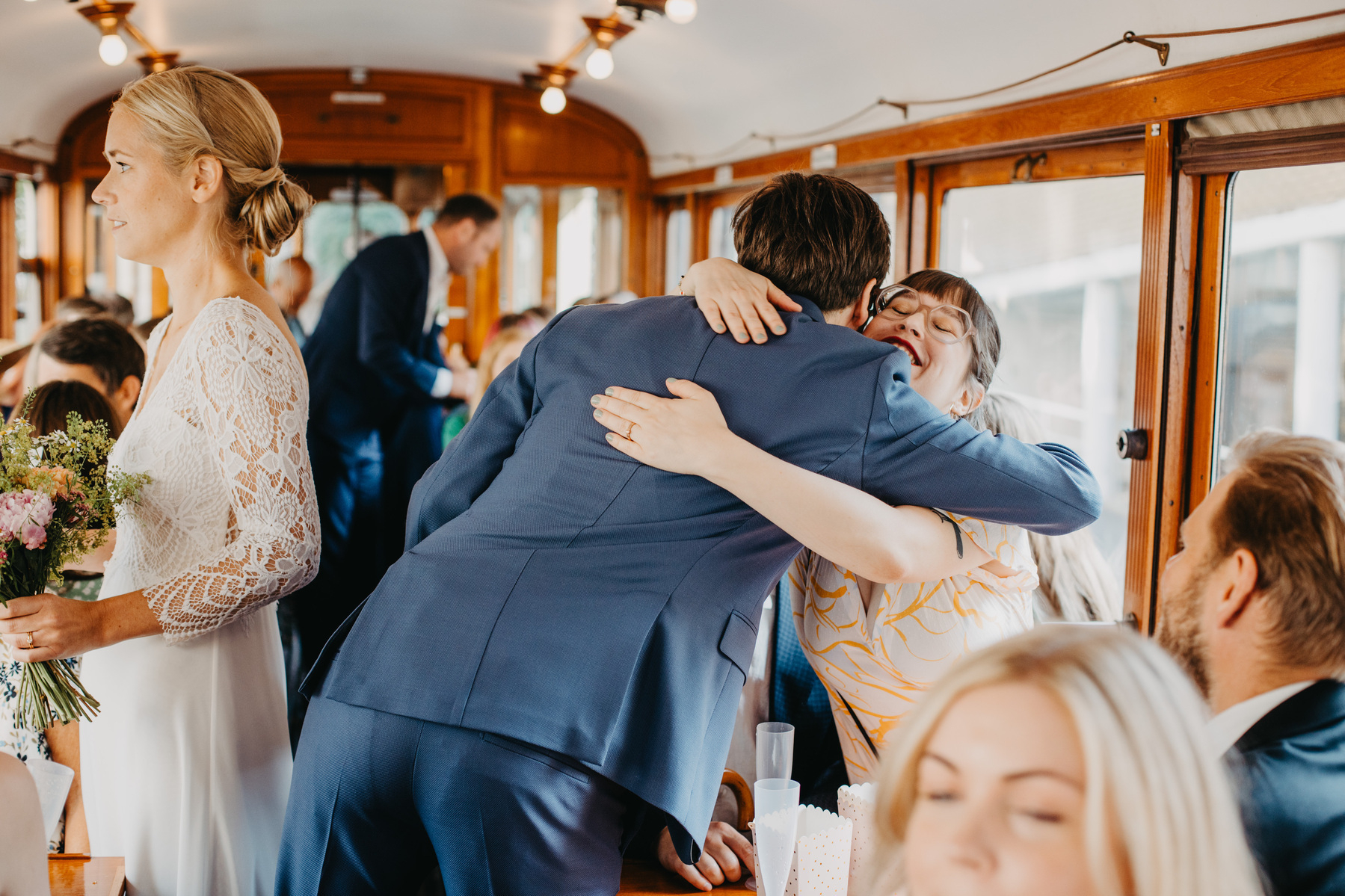 Auto-generated description: A woman dressed in white lace holds a bouquet while a man in a blue suit hugs another woman dressed in yellow and white on a train filled with people.