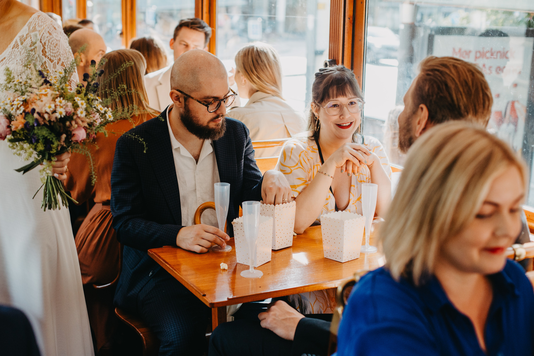 Auto-generated description: Several people are sitting and conversing at a dining table, with one person in a white dress holding a bouquet.