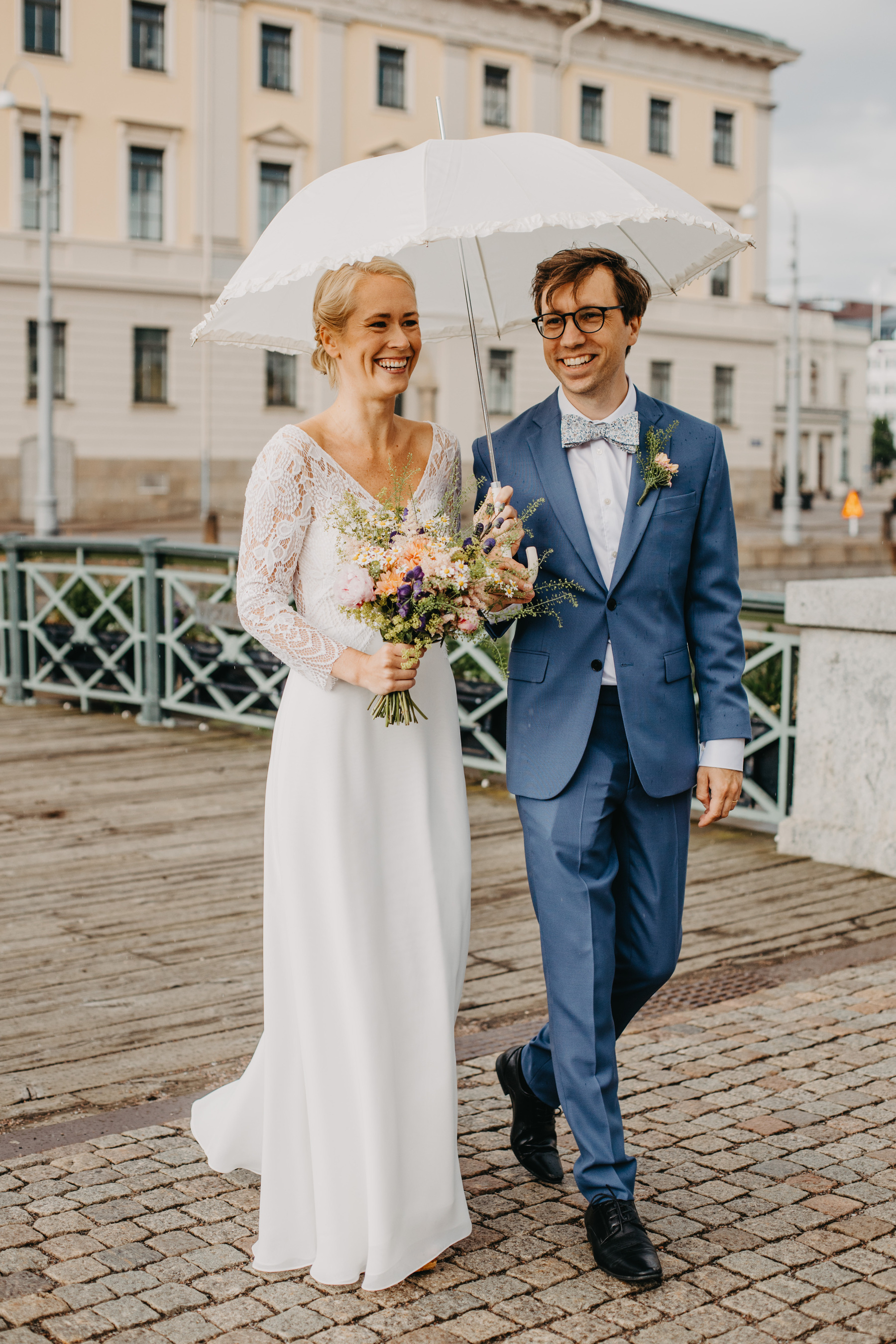 Auto-generated description: A bride and groom, dressed in wedding attire, are walking together outdoors under a white umbrella, smiling joyfully.