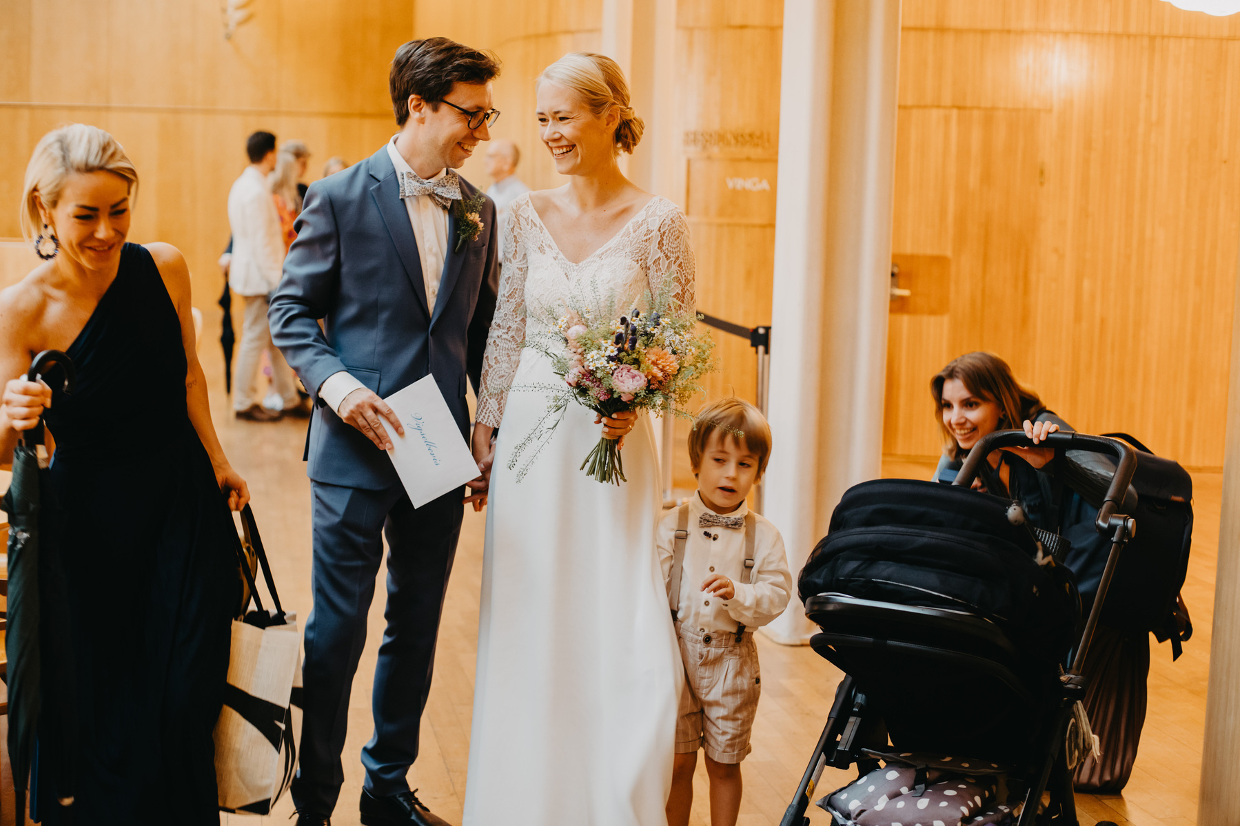 Auto-generated description: A wedding couple is smiling and holding hands while standing with a young boy and two women, one of whom is pushing a stroller.