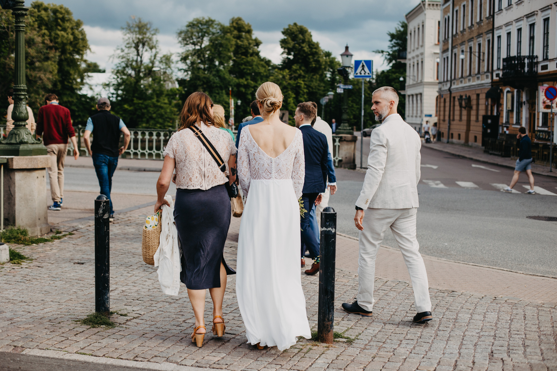 Auto-generated description: A group of people, with a woman in a white dress and another in a lace top, walk along a cobblestone path in an urban area with parked cars and trees in the background.