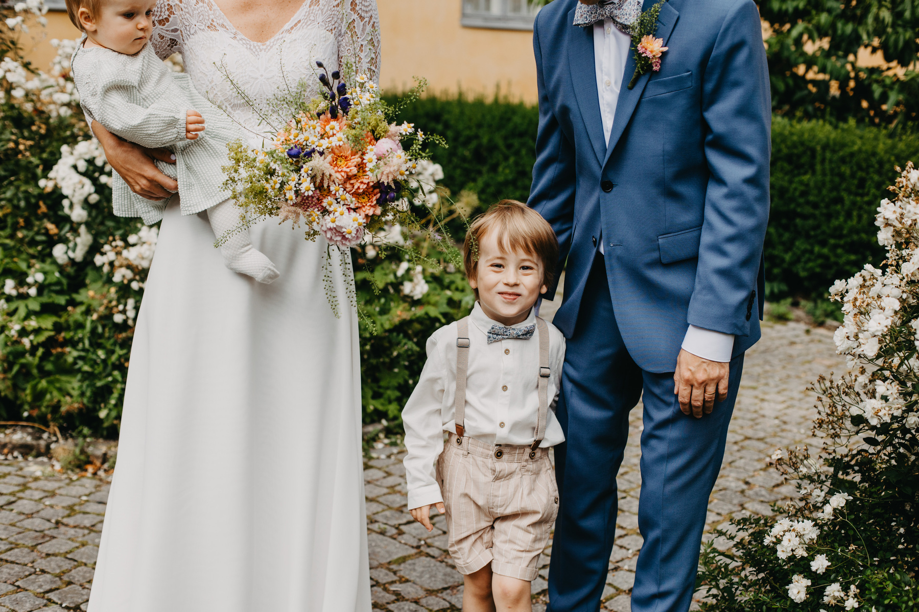 Auto-generated description: A couple dressed in wedding attire stands outdoors with a toddler and a baby, surrounded by greenery and flowers.