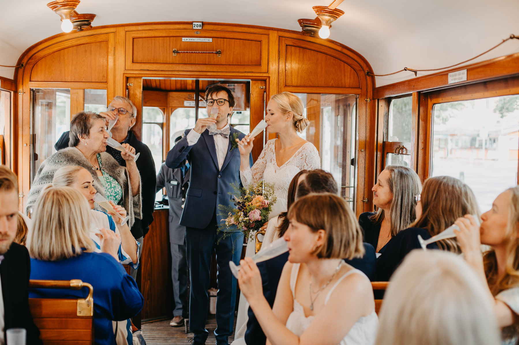 Auto-generated description: A newlywed couple is standing in a vintage trolley car, surrounded by seated guests who are toasting with drinks.