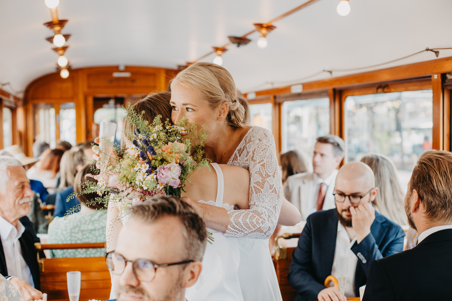 Auto-generated description: A celebration or gathering inside a decorated, well-lit tram with people dressed in formal attire, centered around two women embracing.