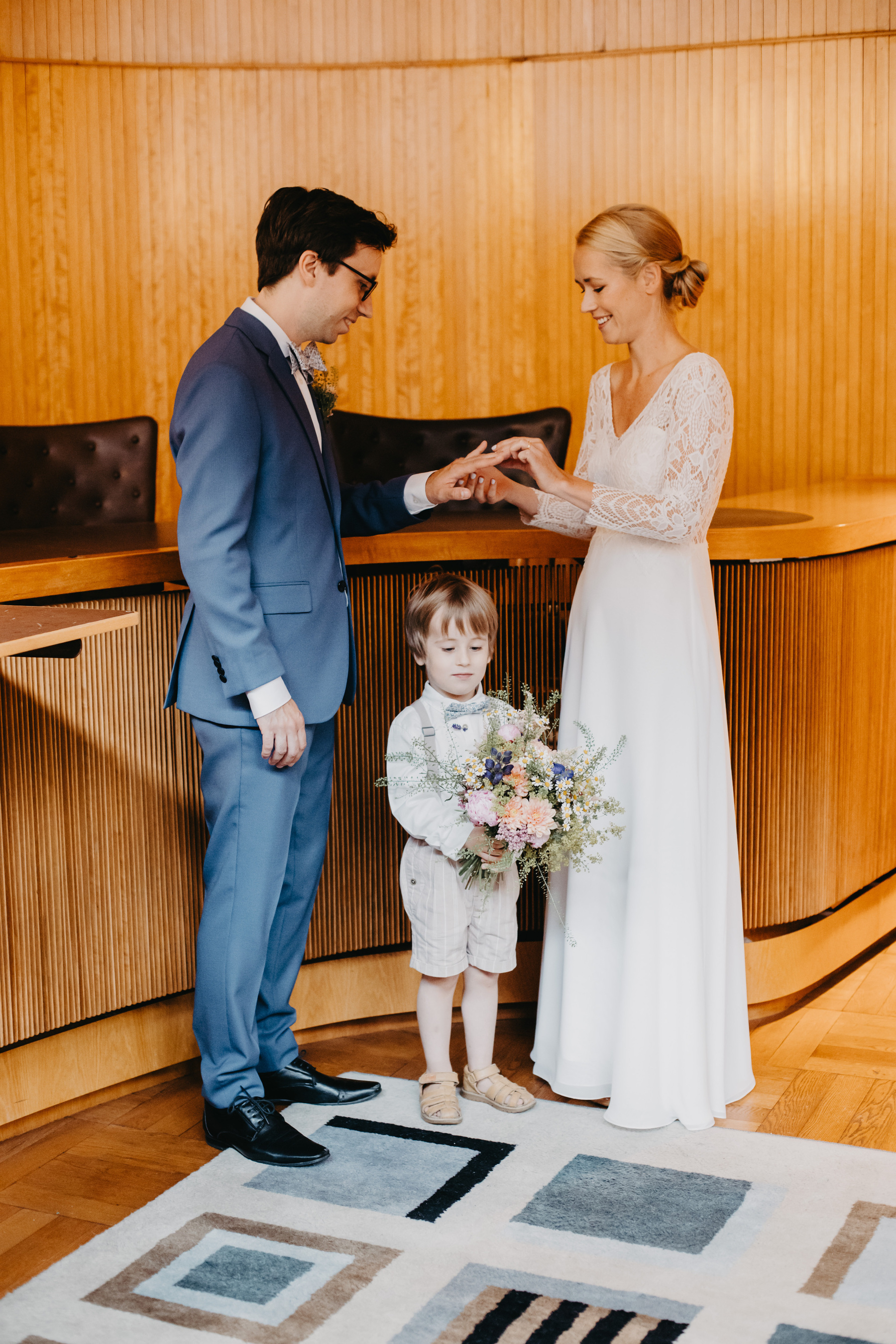 Auto-generated description: A couple dressed in wedding attire is exchanging rings, while a young child holding a bouquet stands between them.