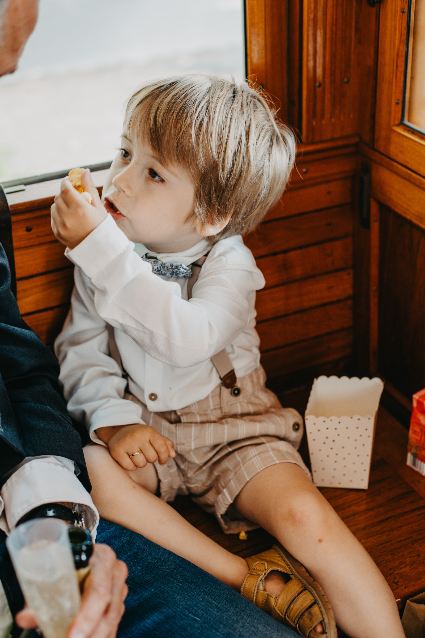 Auto-generated description: A young child with light-colored hair is sitting on a wooden seat near a window, holding a piece of food near their mouth, with an open container of food beside them.