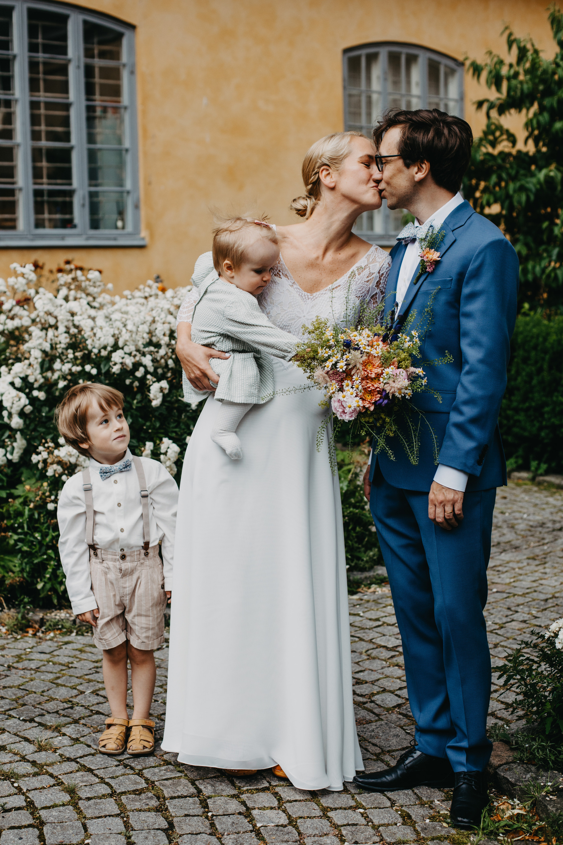 Auto-generated description: A couple is kissing while holding a baby, with a young child standing beside them on a cobblestone path in front of a house with blooming flowers.