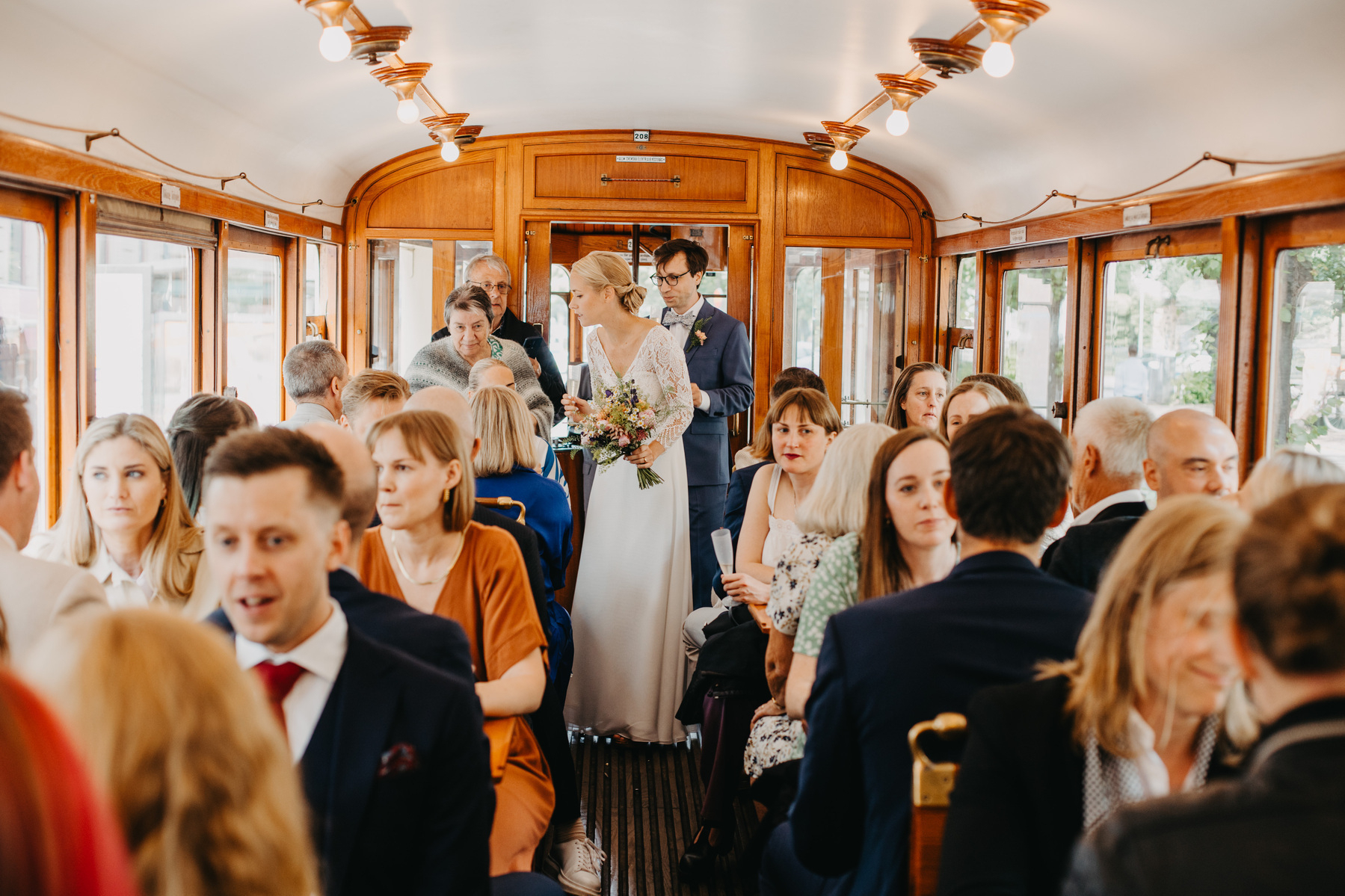 Auto-generated description: A bride and groom stand together inside a crowded, vintage-style tram surrounded by seated guests.