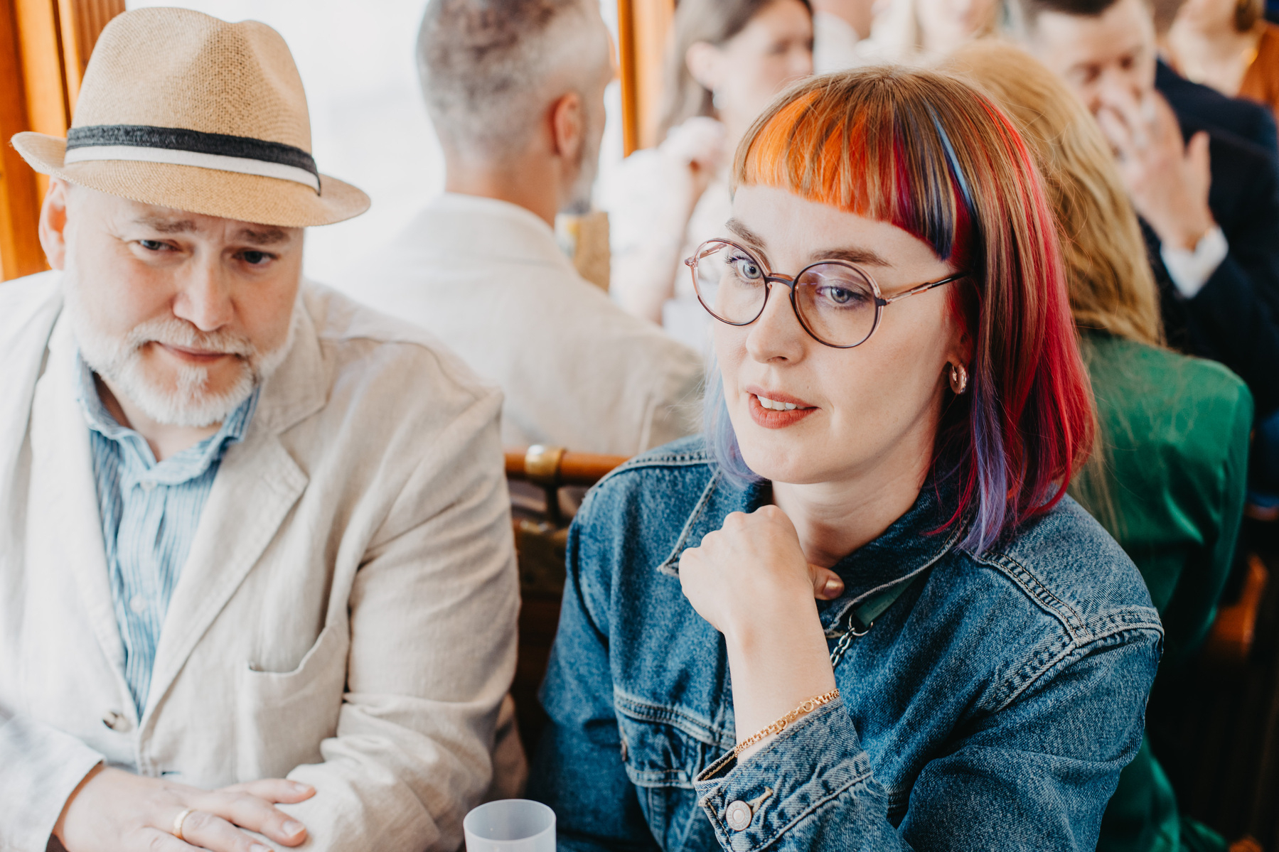 Auto-generated description: A man in a light blazer and hat sits beside a woman with colorful hair and glasses in a crowded indoor setting.