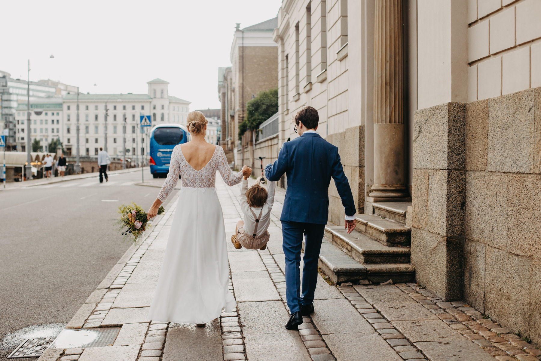 Auto-generated description: A bride and groom are walking down a city street, each holding one of their young child’s hands and lifting the child into the air.