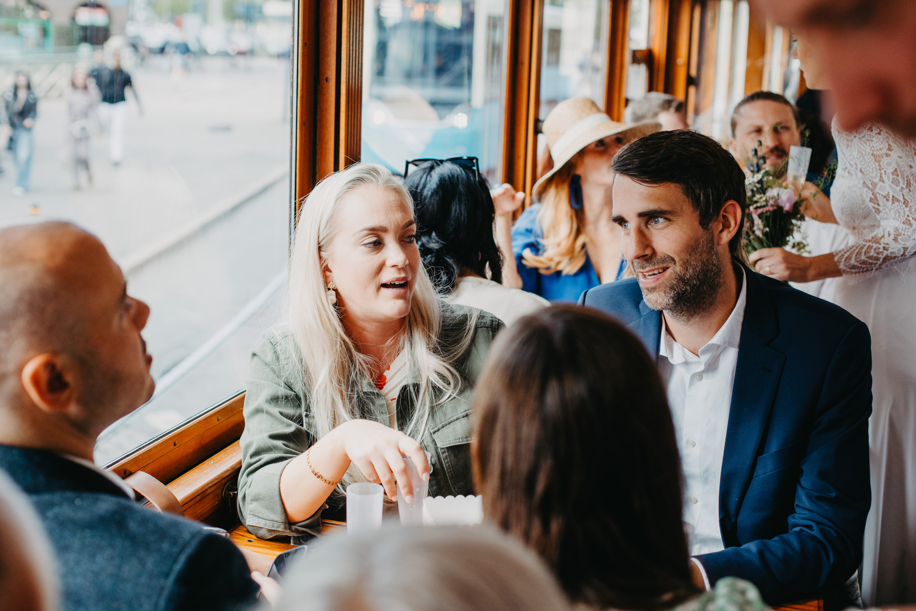 Auto-generated description: People are seated and conversing on what appears to be a moving vehicle with large windows, possibly a tram or train.