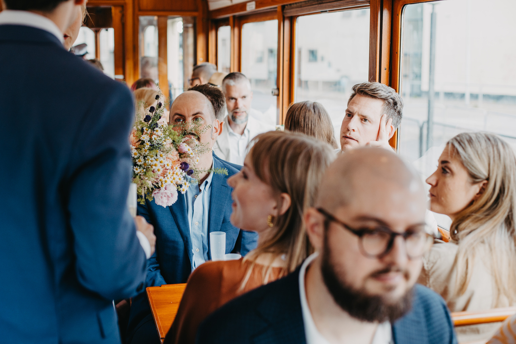 Auto-generated description: People are seated inside a tram or trolley car, engaging in conversation and holding flowers.