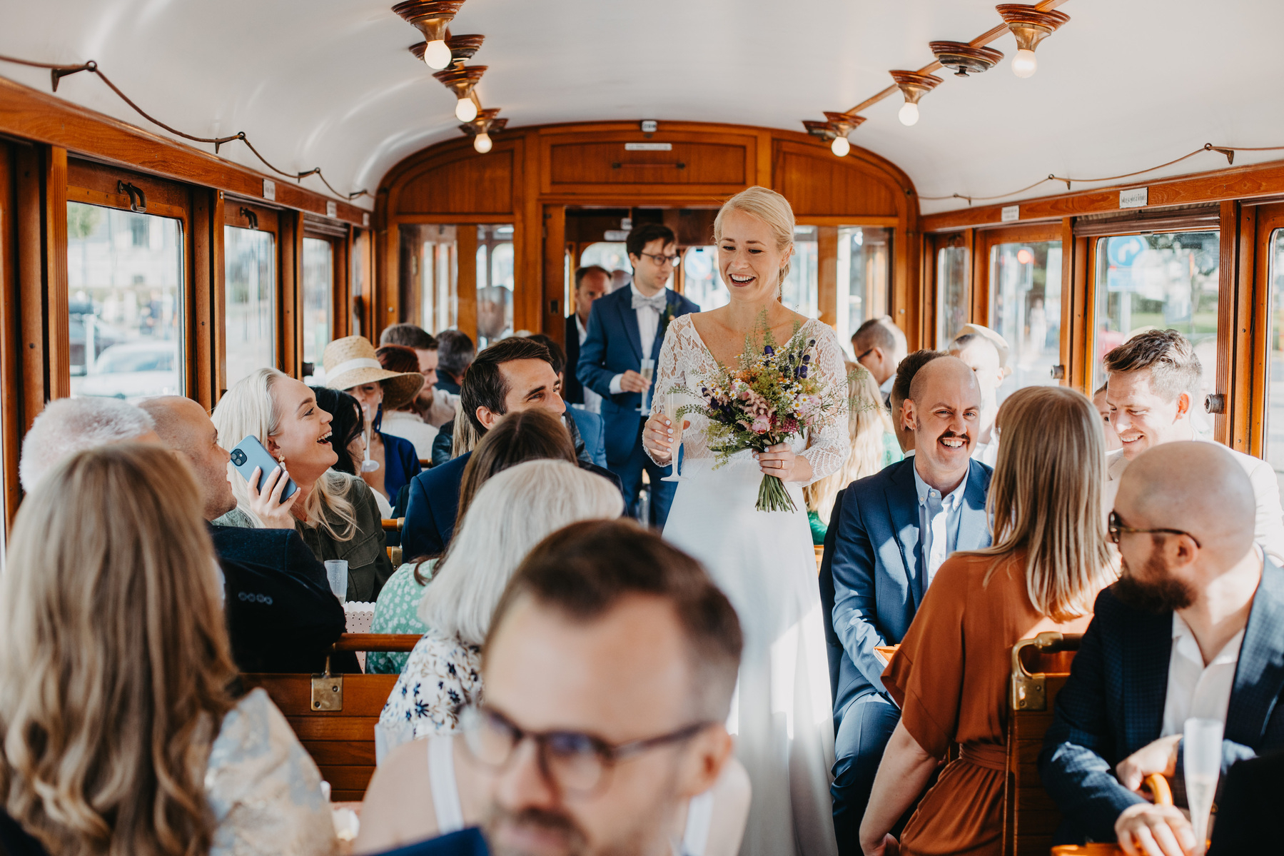 Auto-generated description: A smiling bride holding a bouquet walks down the aisle of a vintage-style tram filled with seated guests.