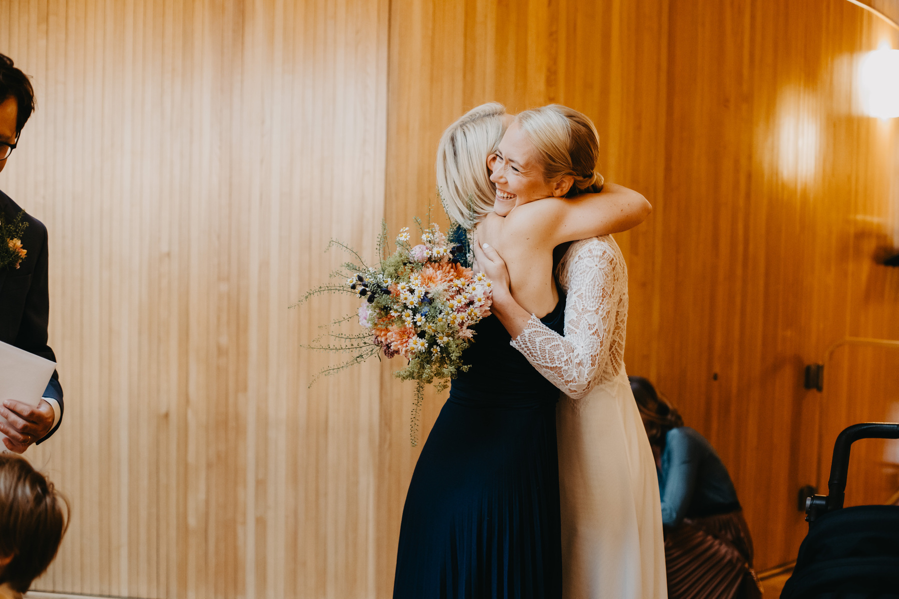 Auto-generated description: Two women, one in a white dress holding a bouquet and the other in a dark dress, are joyfully hugging in a warmly lit wooden room.