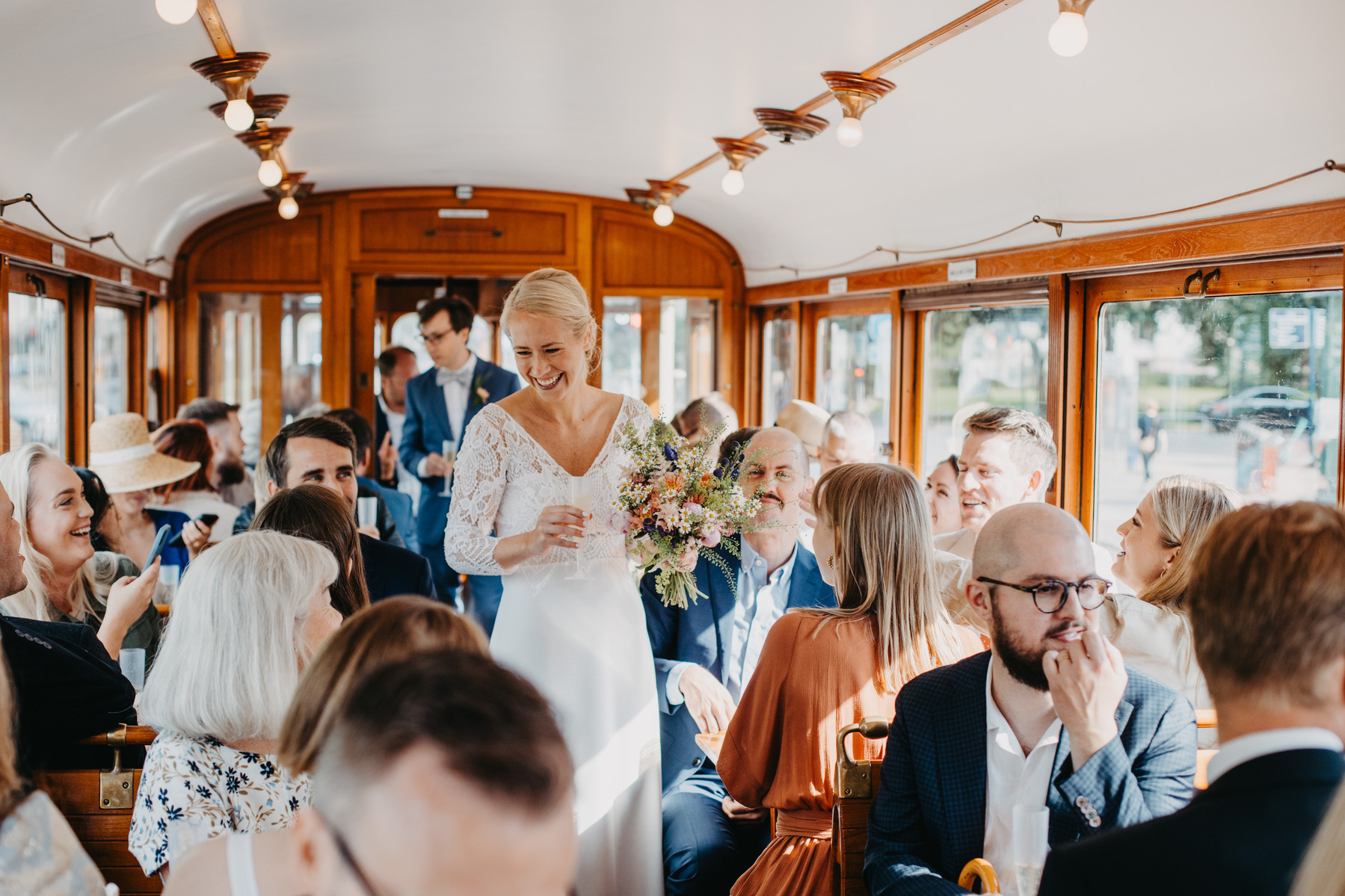 Auto-generated description: A bride, holding a bouquet and wearing a white dress, smiles and interacts with guests inside a charming, vintage tram decorated with warm wooden accents and lights.