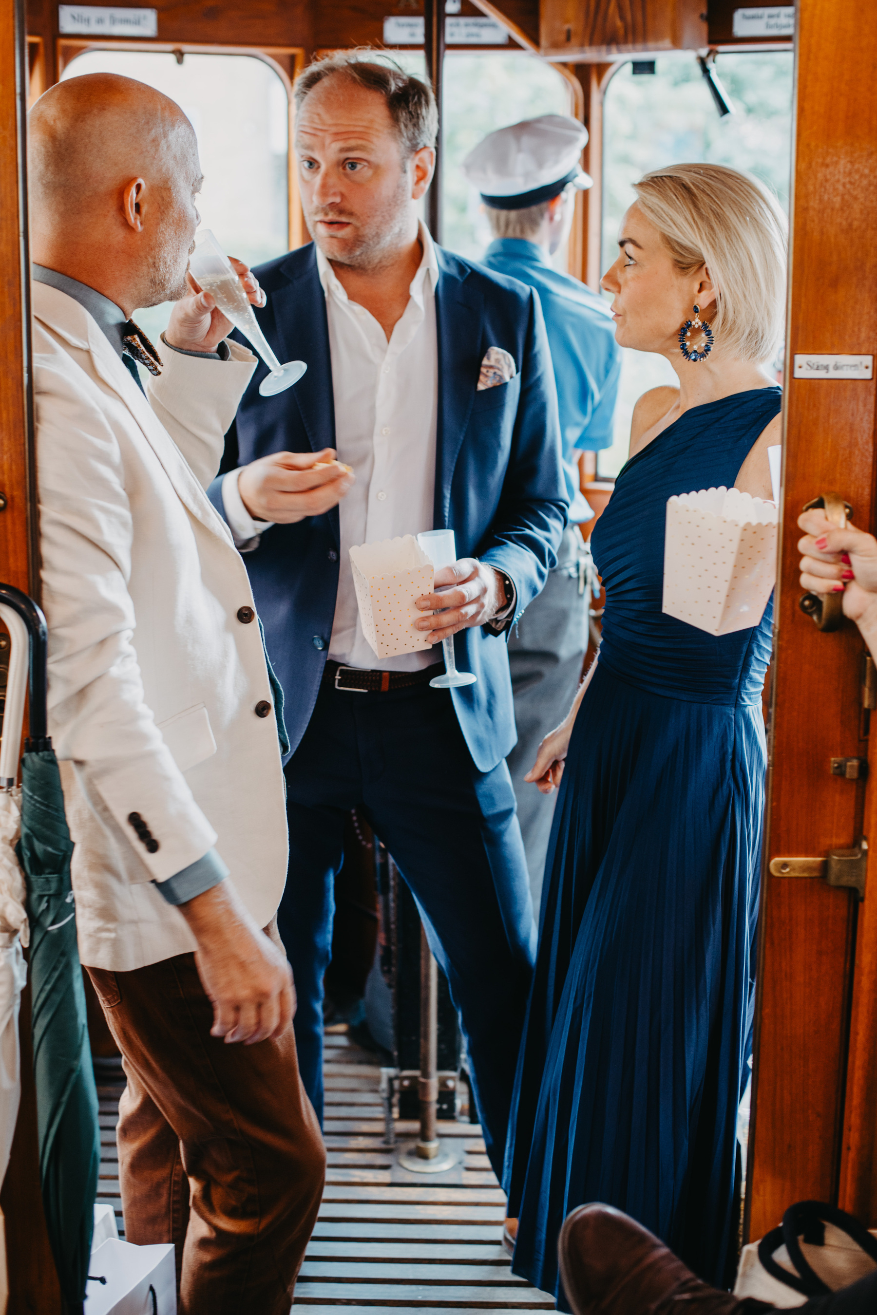 Auto-generated description: Three well-dressed individuals are conversing and enjoying drinks on a tram.