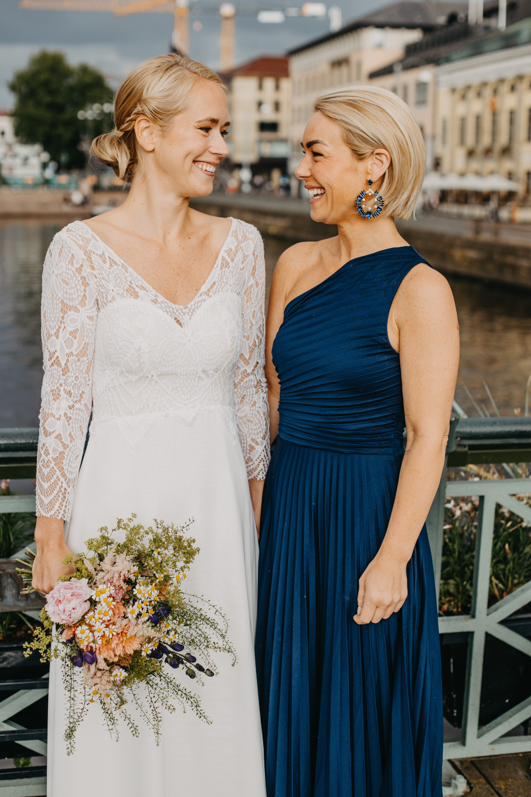 Auto-generated description: Two women in elegant attire, one wearing a white lace gown holding a bouquet and the other in a blue one-shoulder dress, smile at each other while standing outdoors on a bridge.