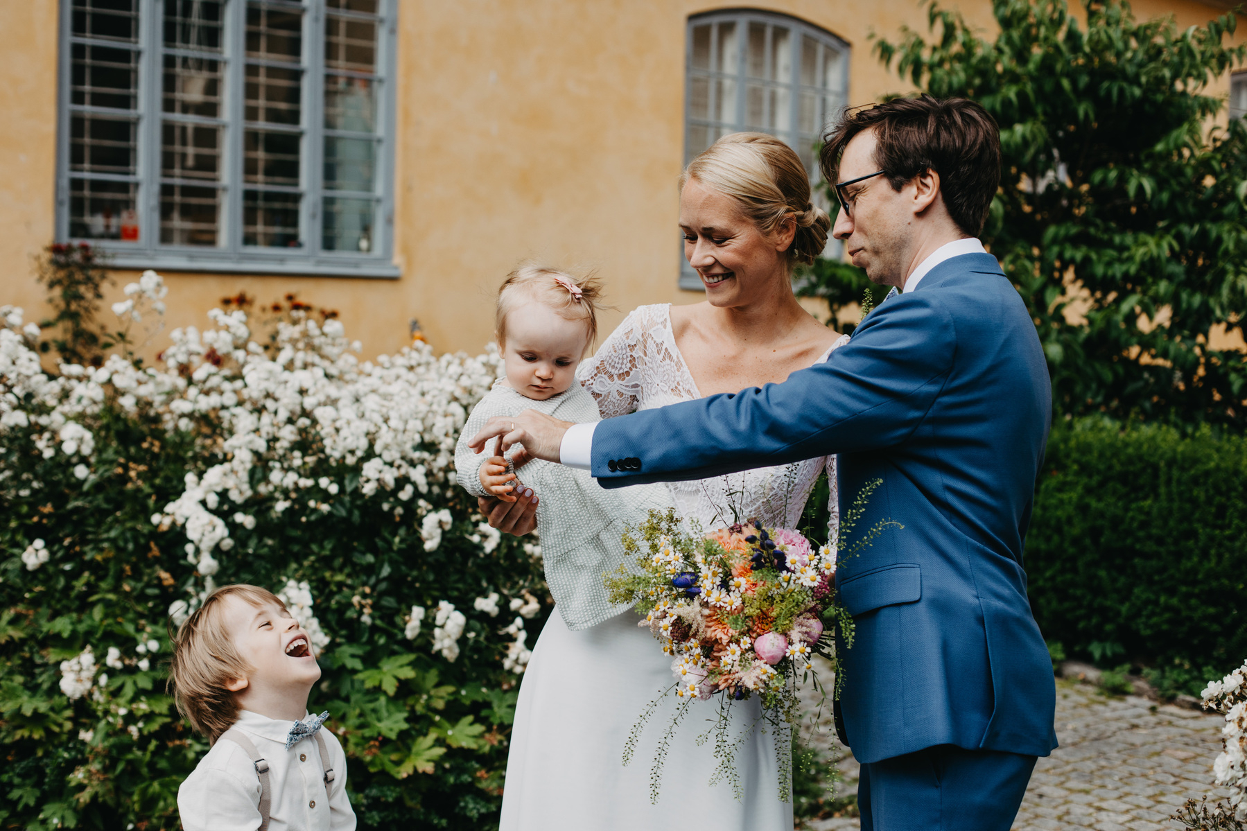 Auto-generated description: A wedding couple stands happily outdoors, with the groom in a blue suit and the bride in a white dress holding both a bouquet and a baby, while a smiling child stands nearby amidst lush greenery and flowers.