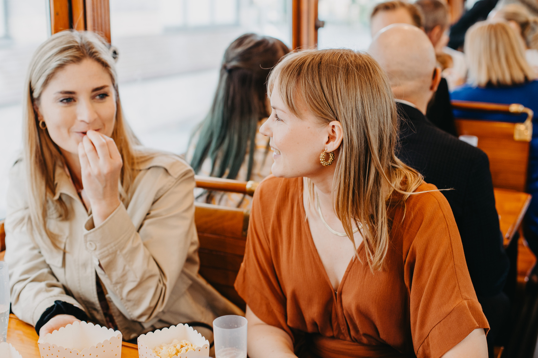 Auto-generated description: Two women are engaged in conversation while sitting at a table in a busy restaurant or cafe, with one of them eating popcorn.