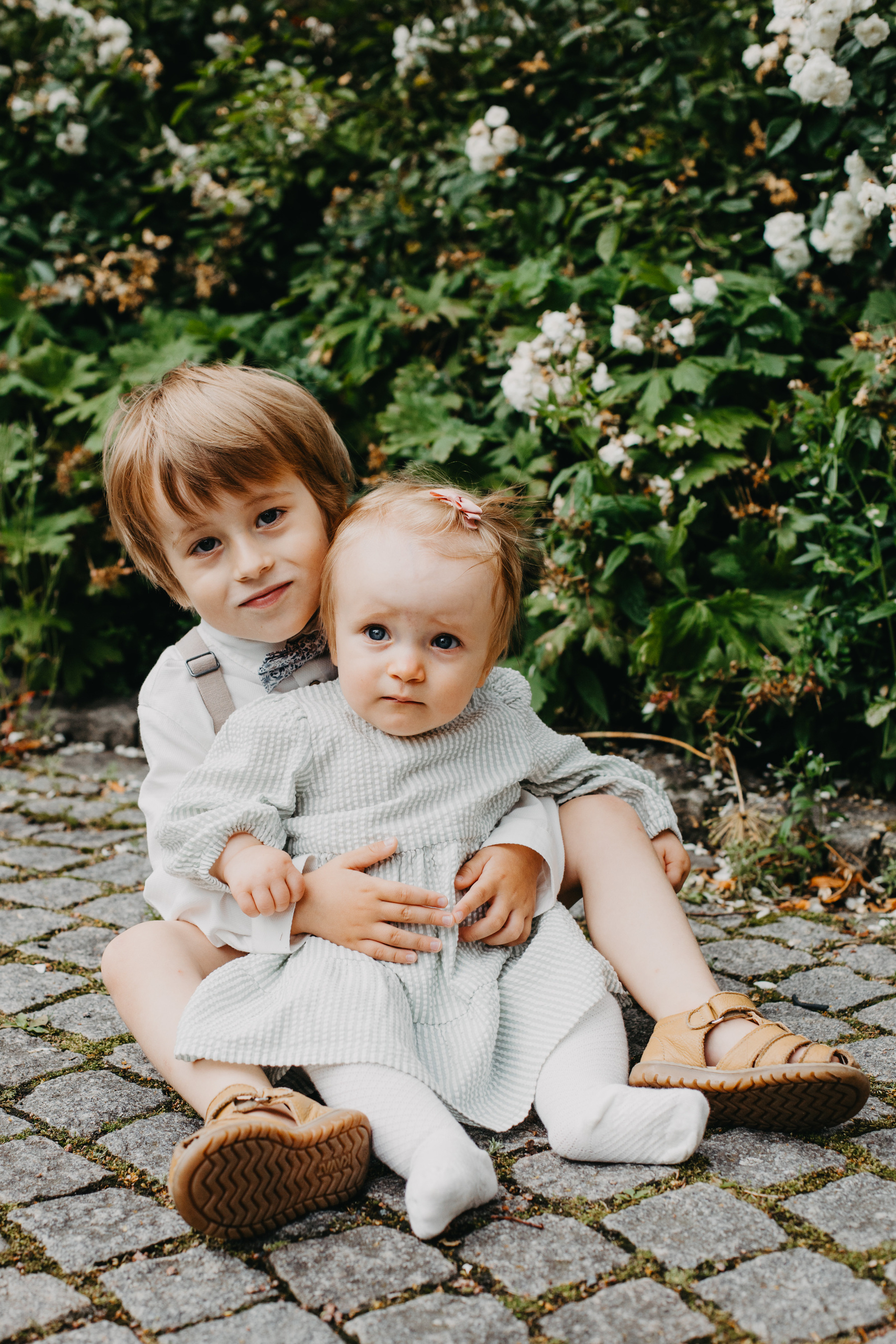 Auto-generated description: Two young children, dressed in light-colored clothes, are sitting on a cobblestone path with greenery and flowers in the background, the older one holding the younger one.