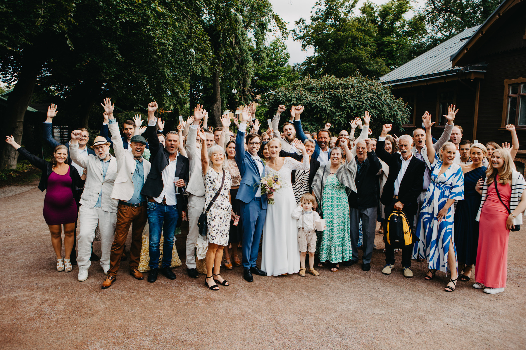 Auto-generated description: A large group of people, dressed in a variety of formal and semi-formal attire, are celebrating outdoors with their hands raised in the air, likely in honor of a wedding.