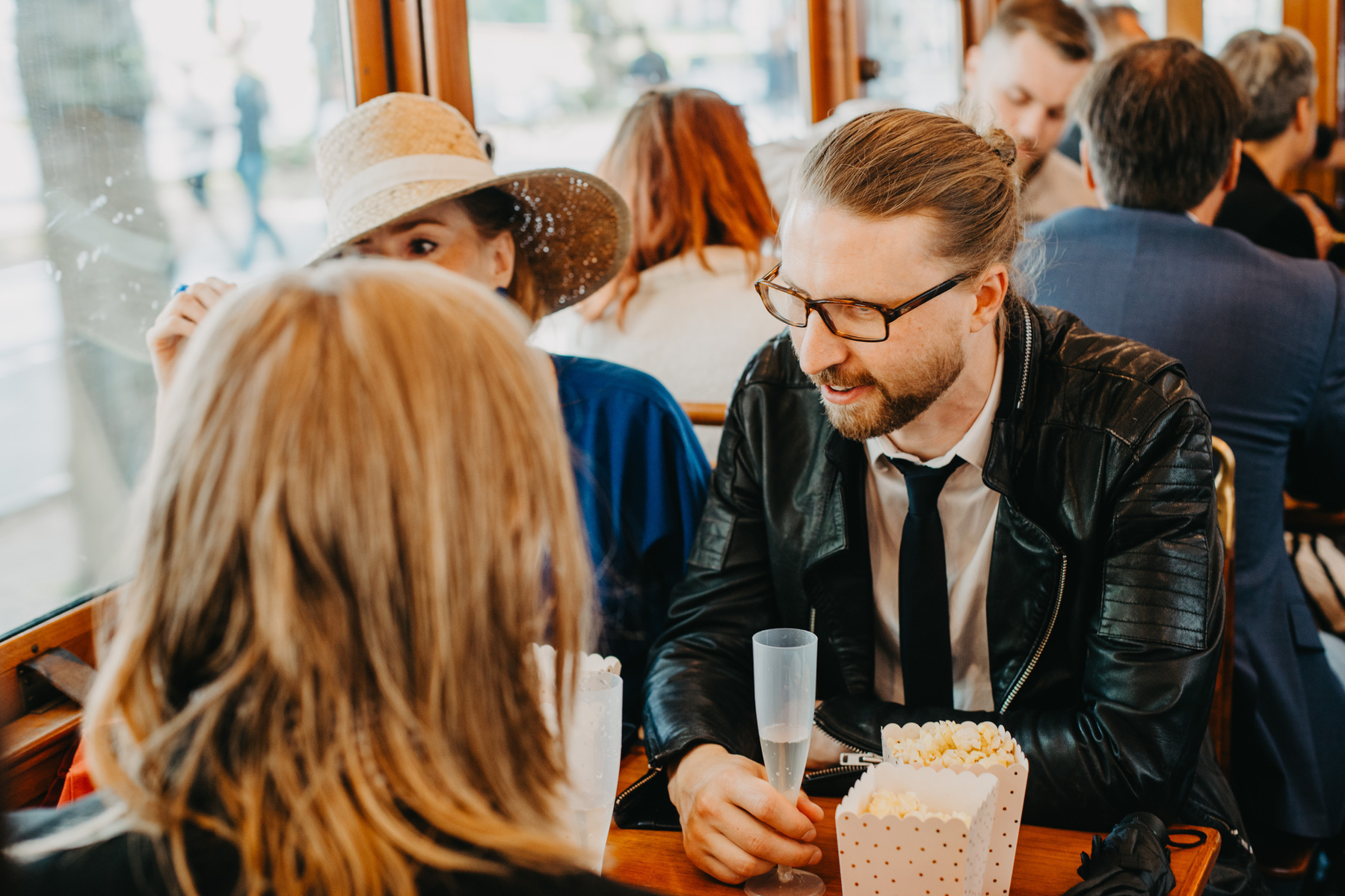 Auto-generated description: A group of people, including a man in a black leather jacket and glasses, are engaged in conversation while sitting at a table with popcorn containers inside a crowded venue.