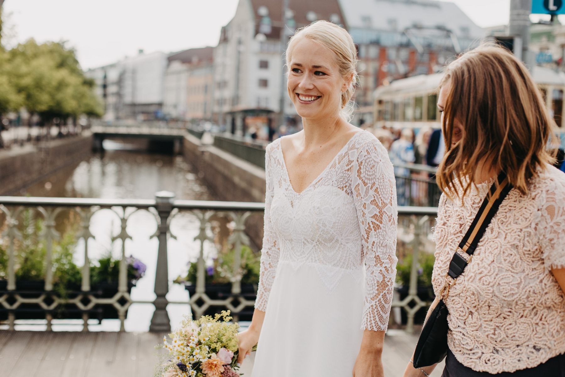 Auto-generated description: A smiling bride in a white lace dress stands on a bridge next to another woman holding a bouquet of flowers, with a canal and city buildings in the background.