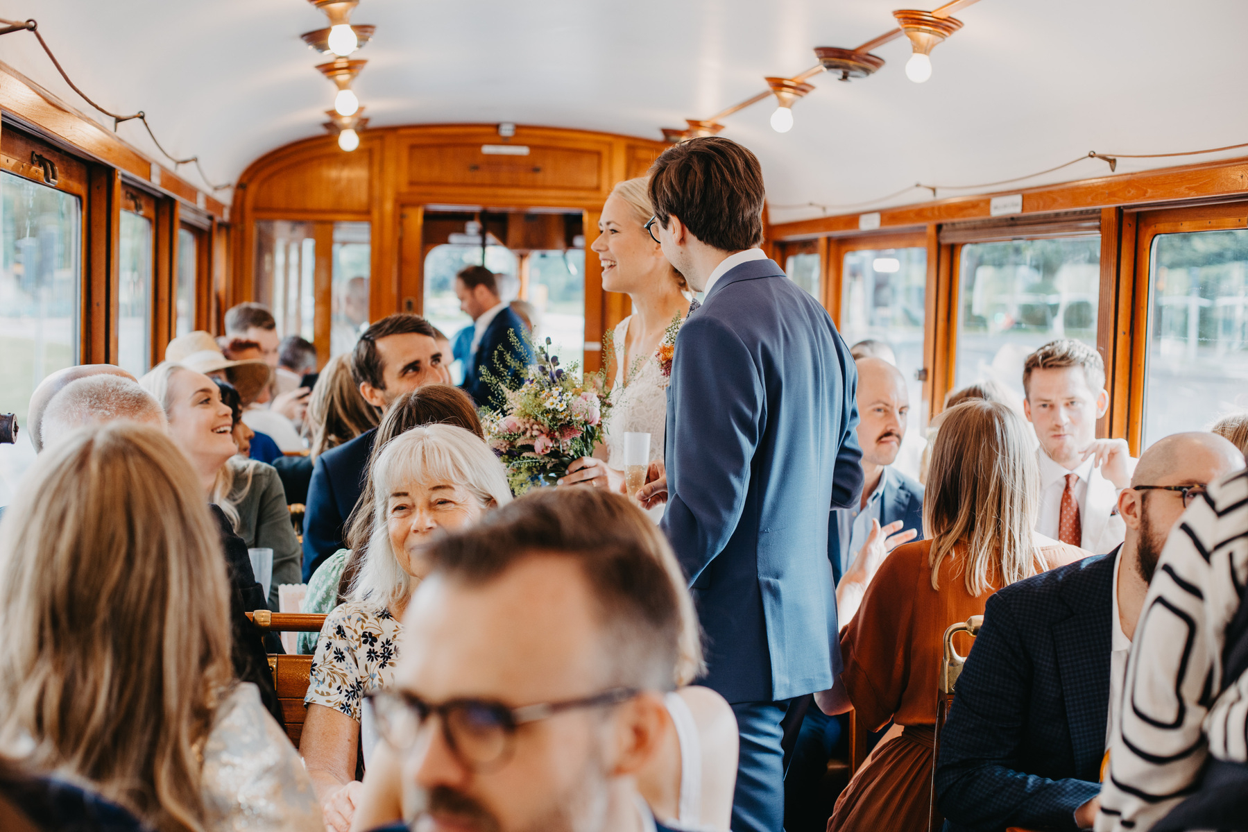 Auto-generated description: A couple dressed in wedding attire stands amidst a group of guests inside a well-lit, wood-paneled trolley car.