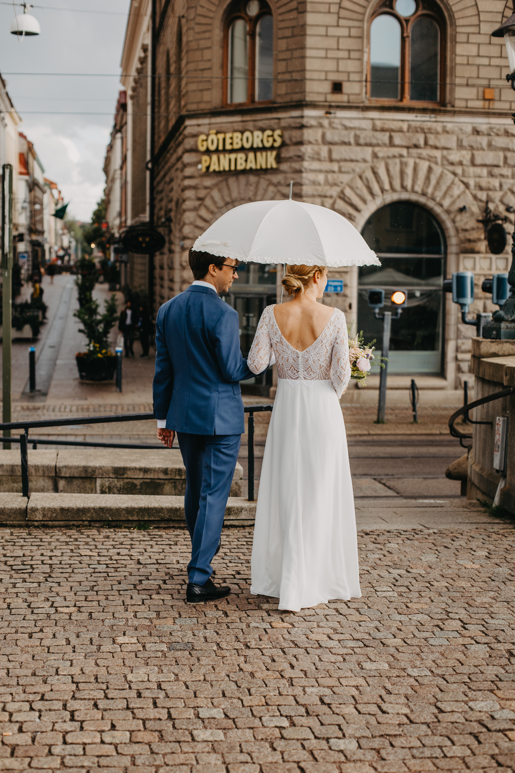 Auto-generated description: A couple dressed in wedding attire walks down a cobblestone street, with the bride holding a white umbrella.