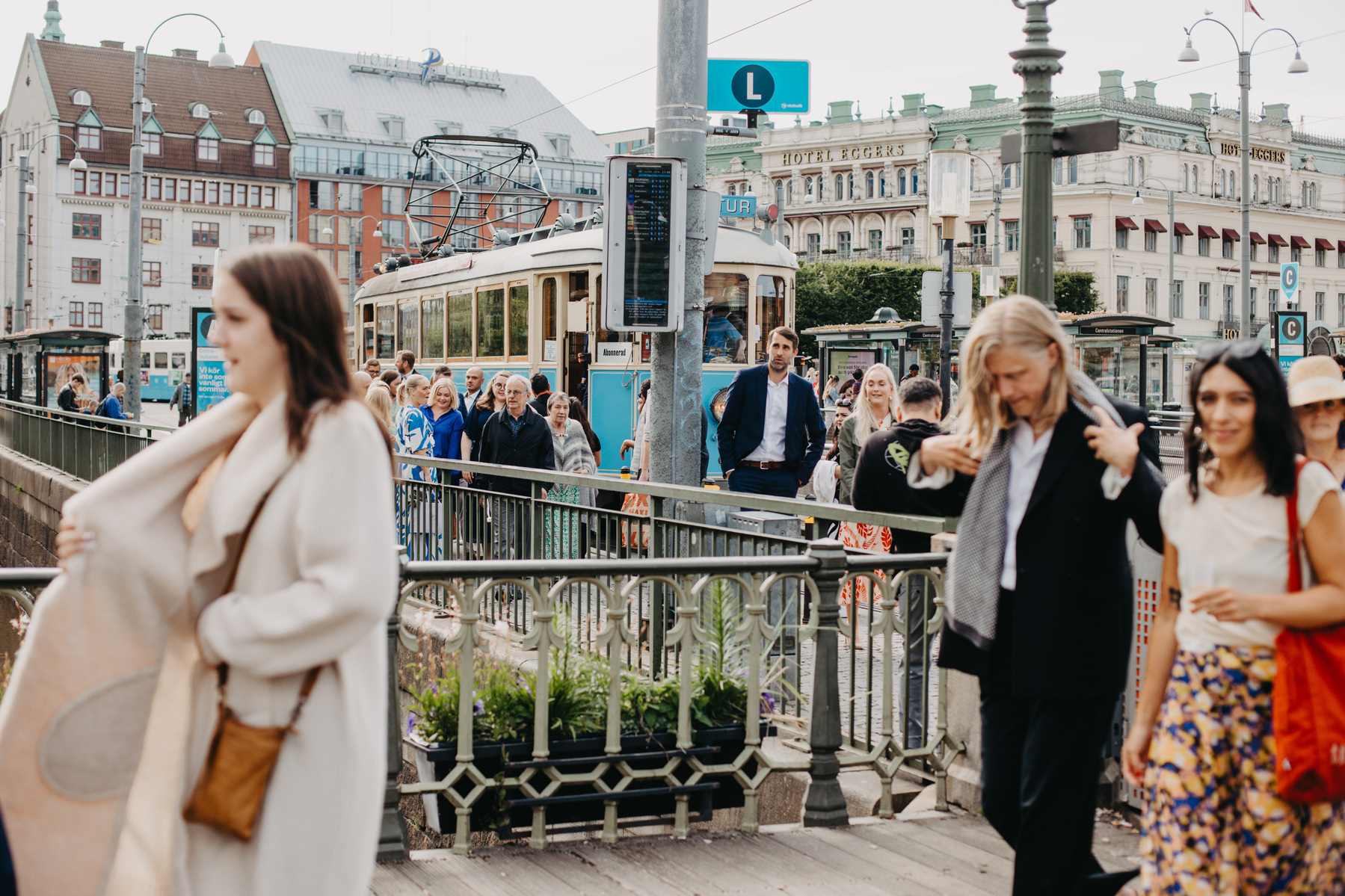 Auto-generated description: A group of people, some walking and some standing, are in an urban setting with an old-fashioned tram and historic buildings in the background.