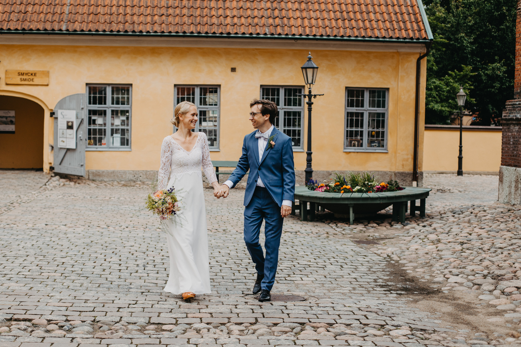 Auto-generated description: A couple is strolling hand-in-hand in a cobblestone courtyard with a quaint yellow building in the background.