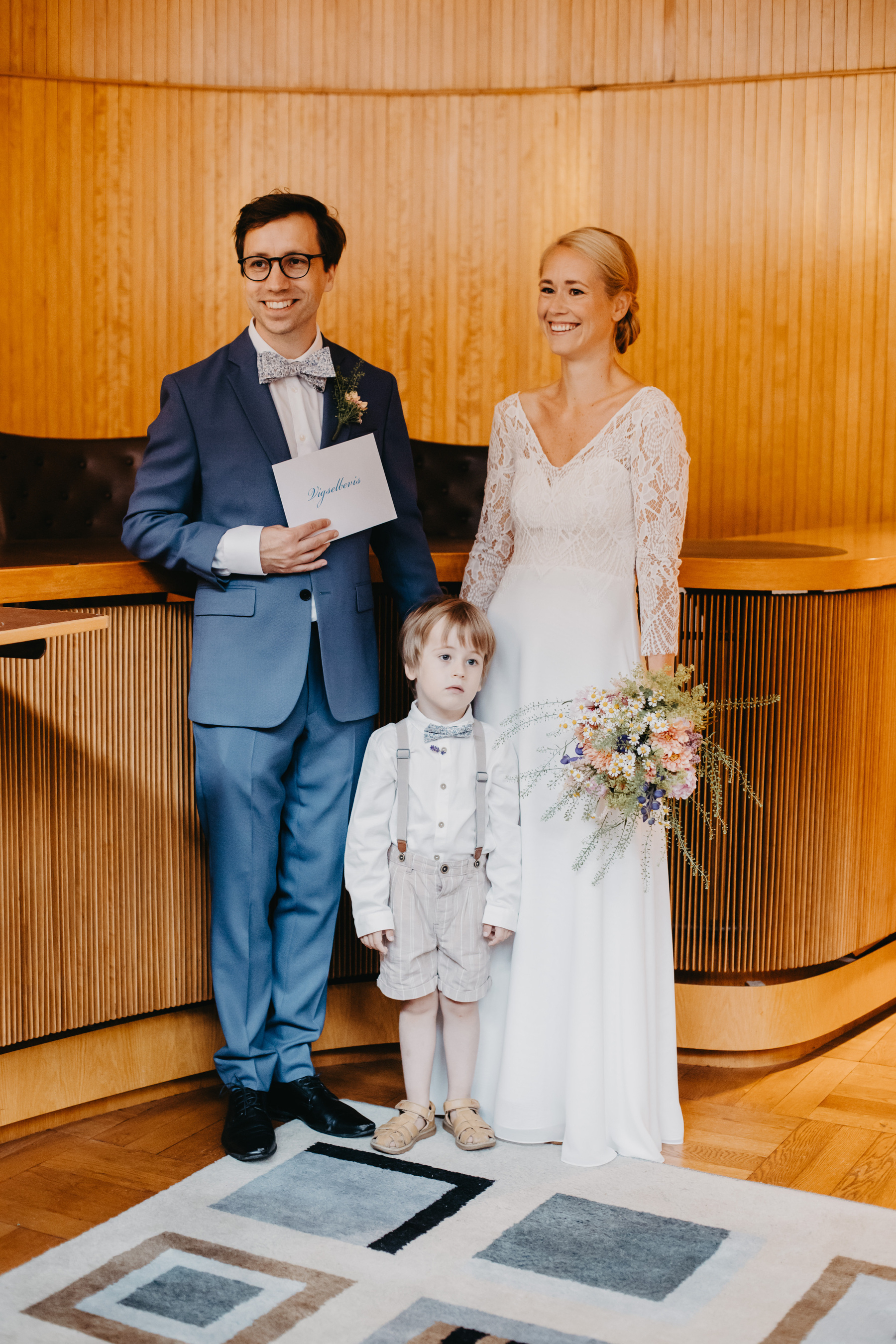 Auto-generated description: A well-dressed couple stands happily in a wood-paneled room, holding a certificate and bouquet, with a young child in front of them.