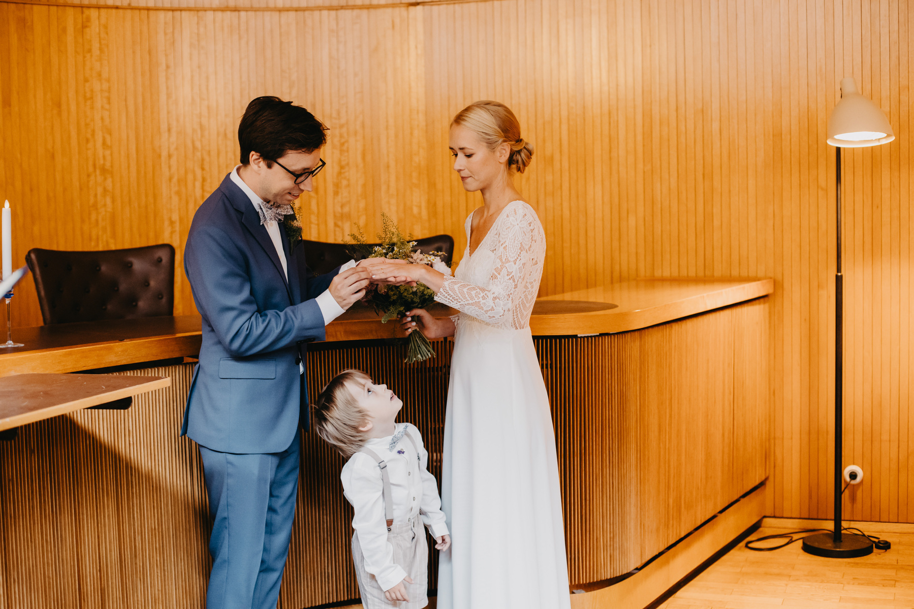 Auto-generated description: A couple is exchanging rings in a wooden-paneled room, with a young child looking up at them.