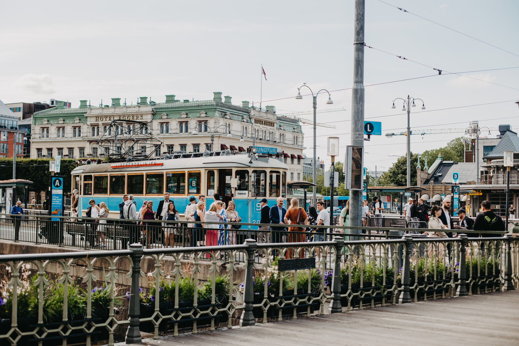 Auto-generated description: A busy tram stop in an urban area with people waiting and a historic building in the background.