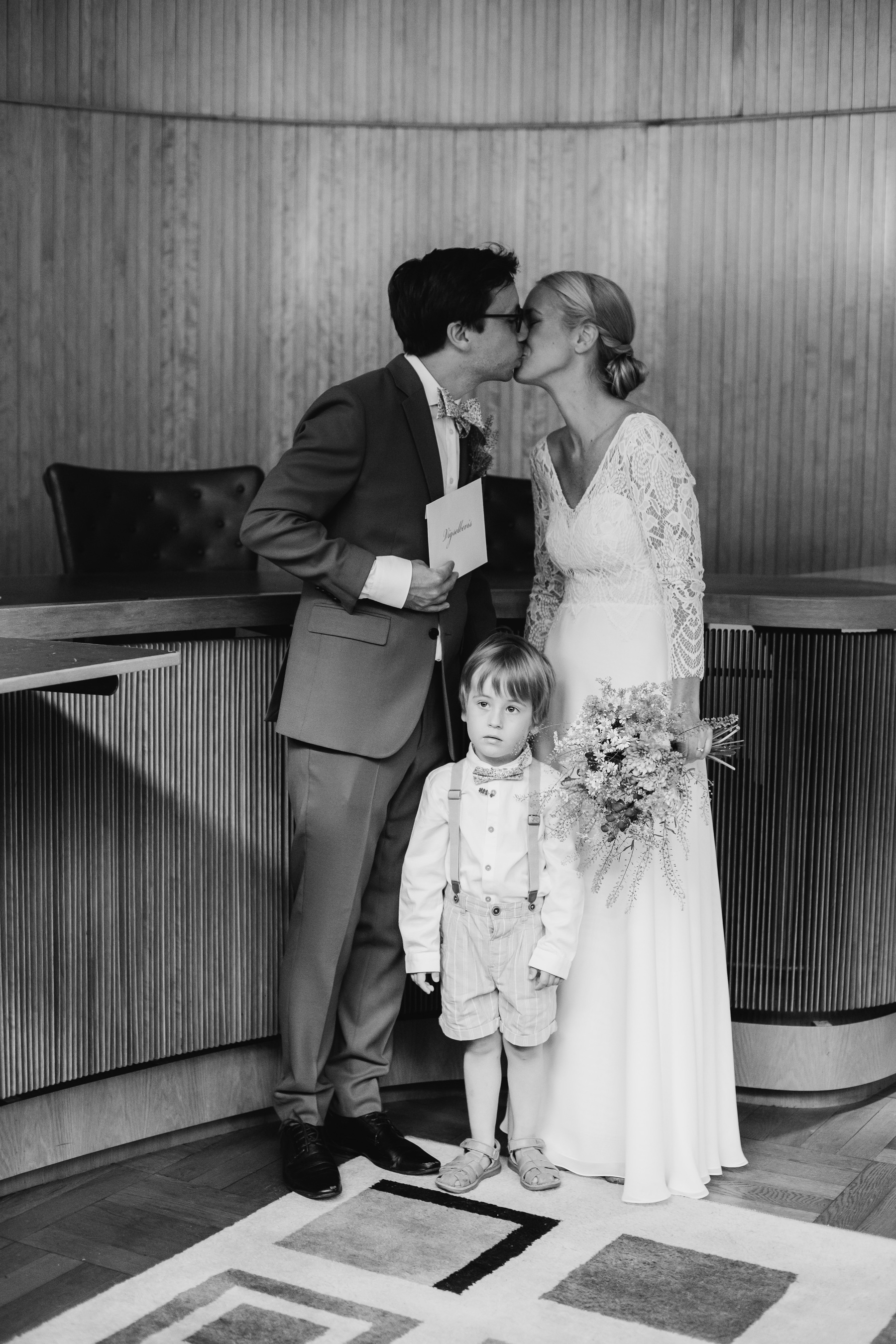 Auto-generated description: A couple in wedding attire shares a kiss while holding their marriage certificate, accompanied by a young child standing in the foreground.