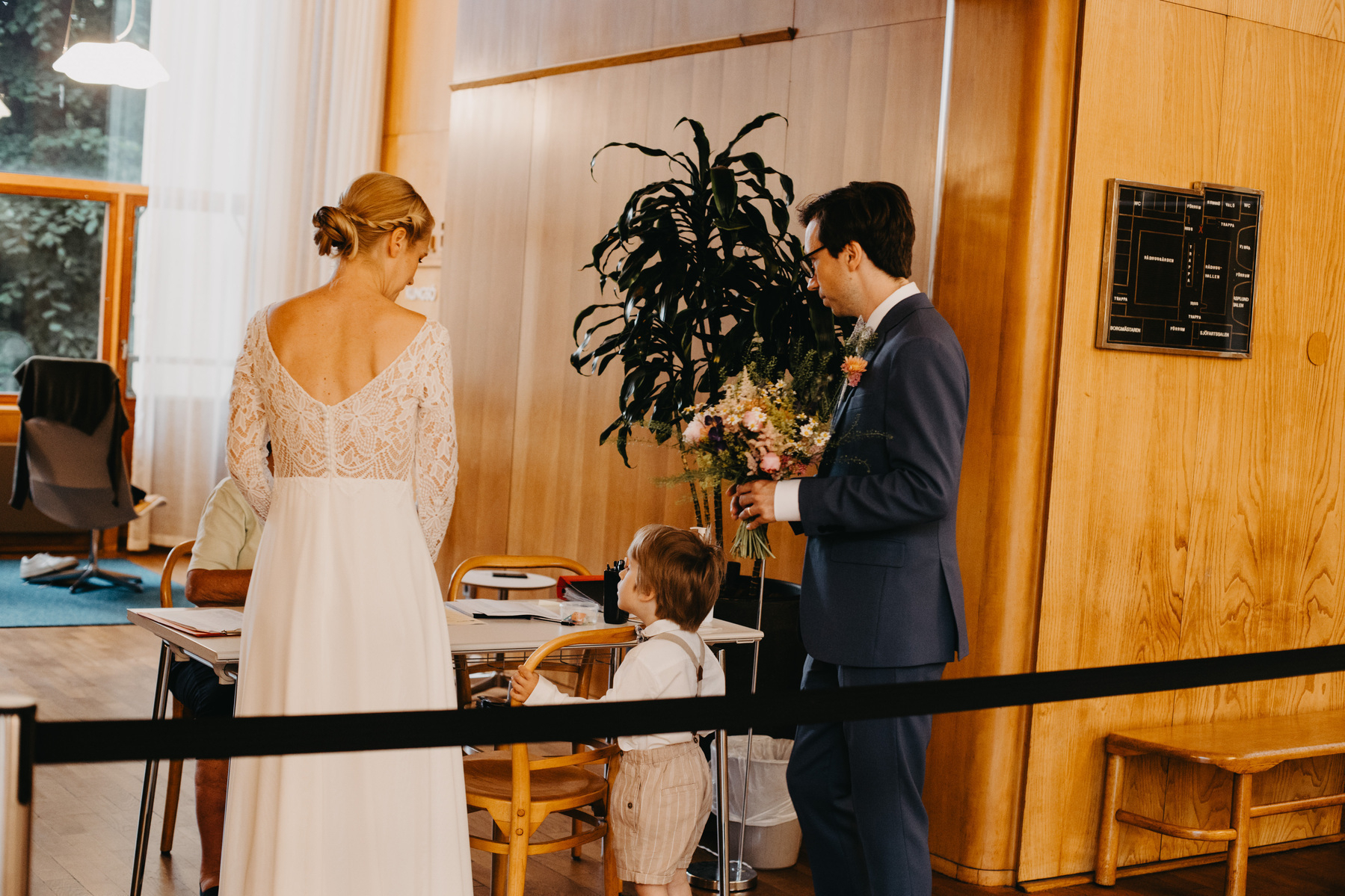 Auto-generated description: A couple dressed in wedding attire, accompanied by a small child, stands at a table inside a wood-paneled room.