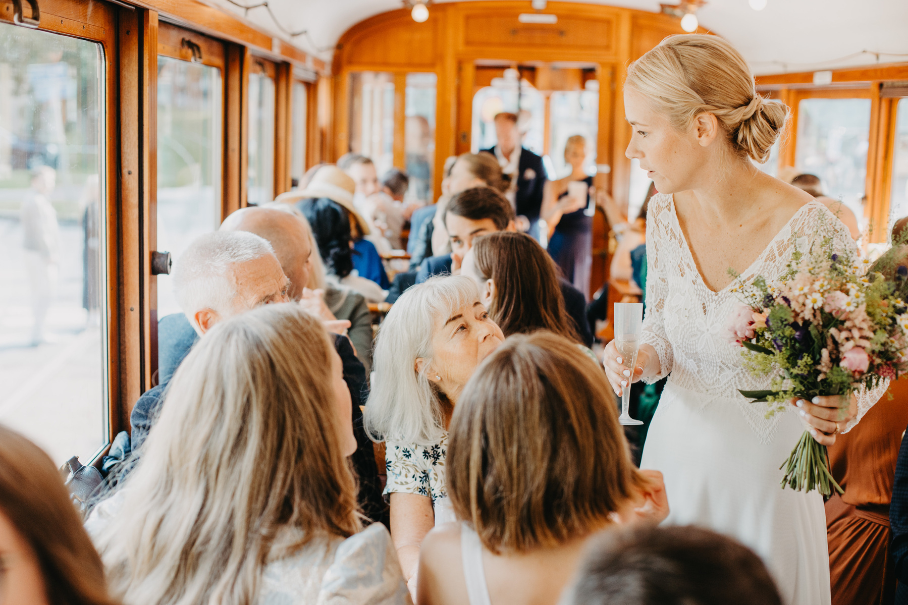 Auto-generated description: A bride holding a bouquet and a glass interacts with guests seated inside a crowded, wood-paneled trolley.