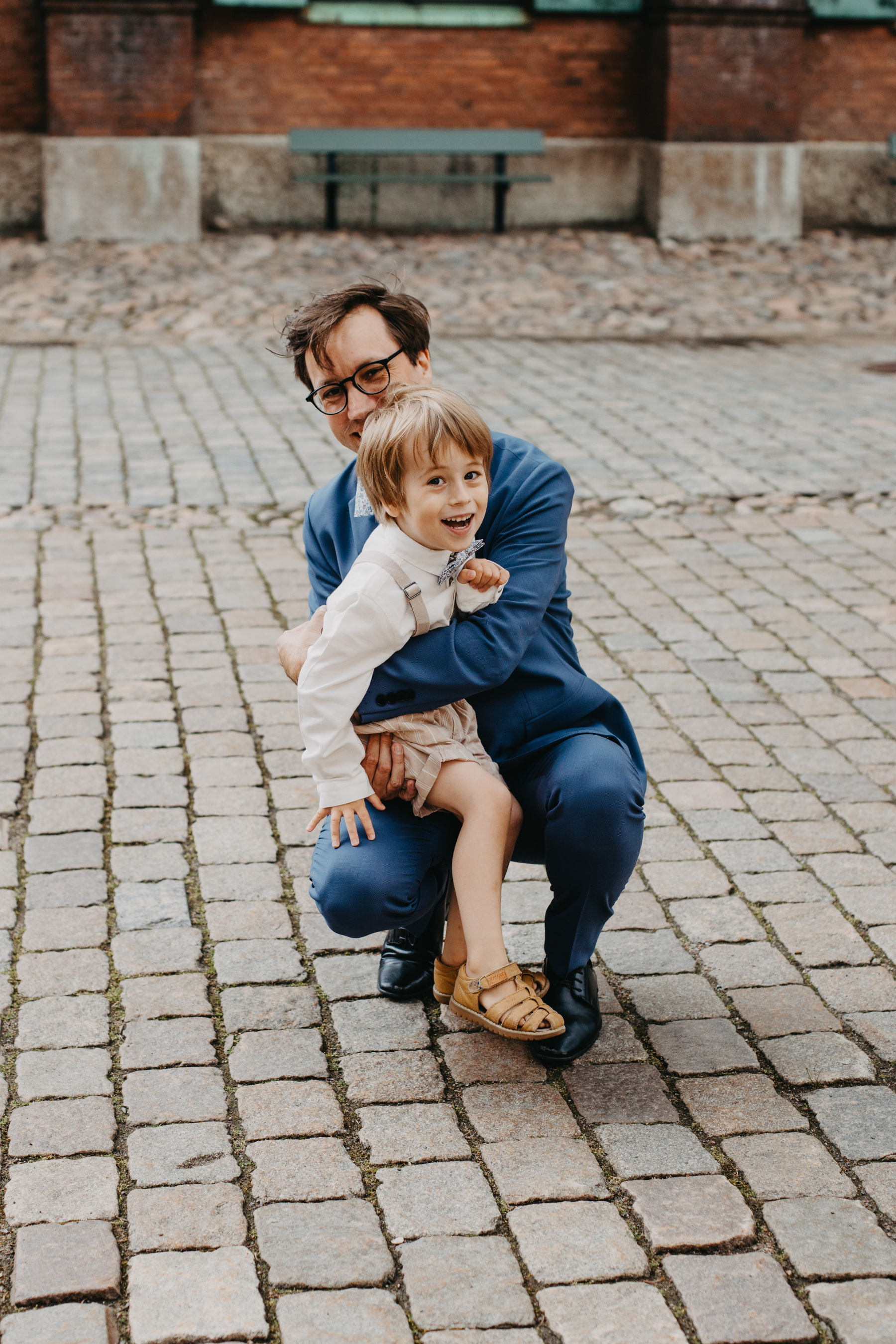 Auto-generated description: A man in a blue suit happily hugs a young child wearing a white shirt and beige shorts while kneeling on a cobblestone path.